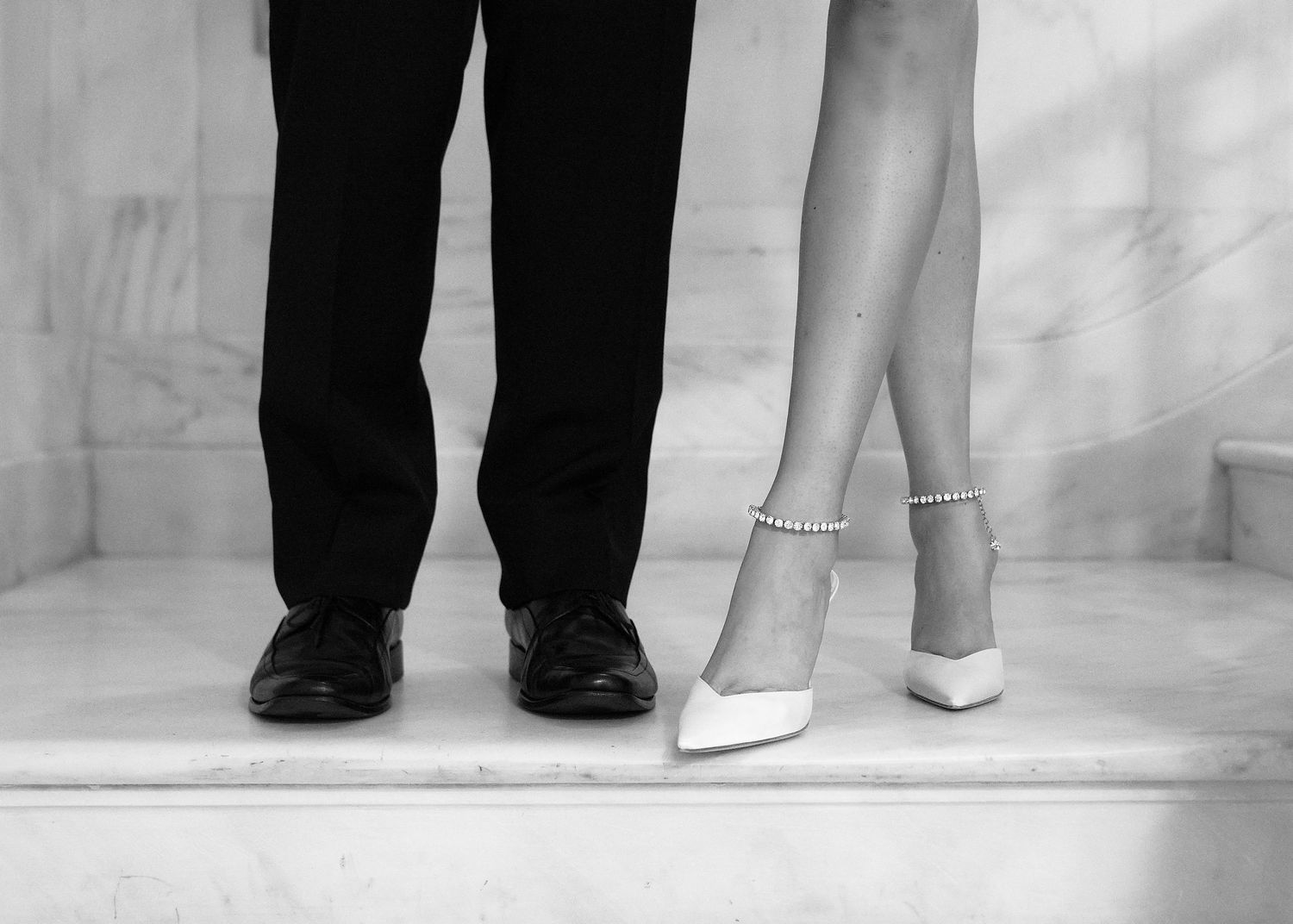 Black and white photo of a couple's feet, man in dress shoes and woman in white heeled pumps with ankle strap.