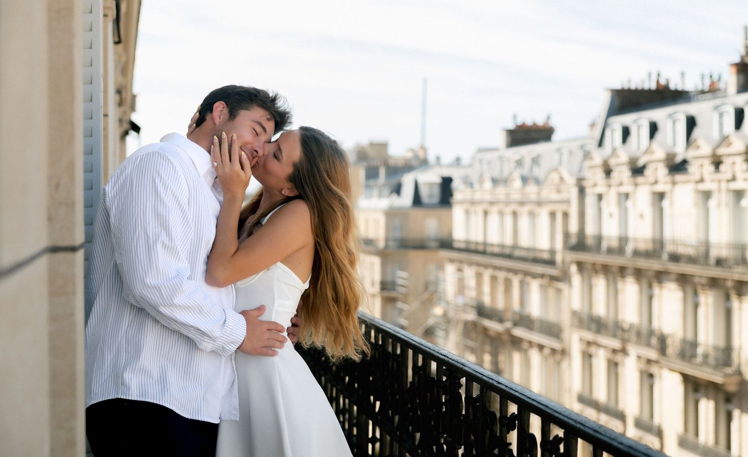 Couple sharing a romantic kiss on a Parisian balcony with classic Haussmann buildings in the background.