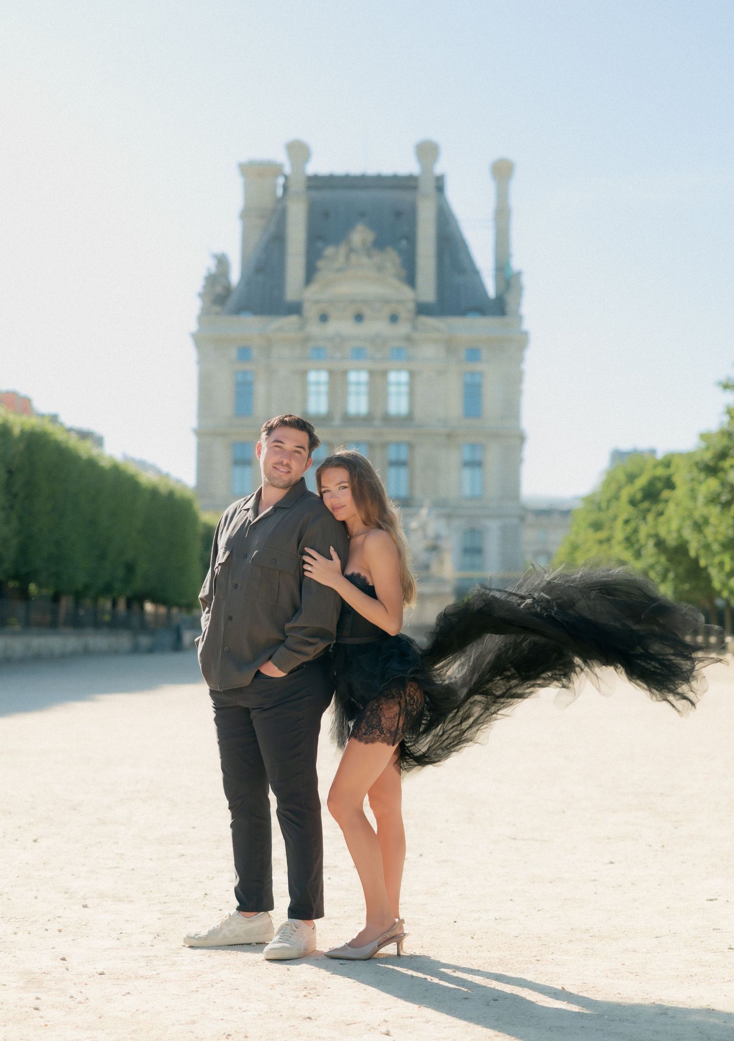 Couple posing in front of the Louvre Palace in Paris, woman in black feathered mini dress, sunny day.