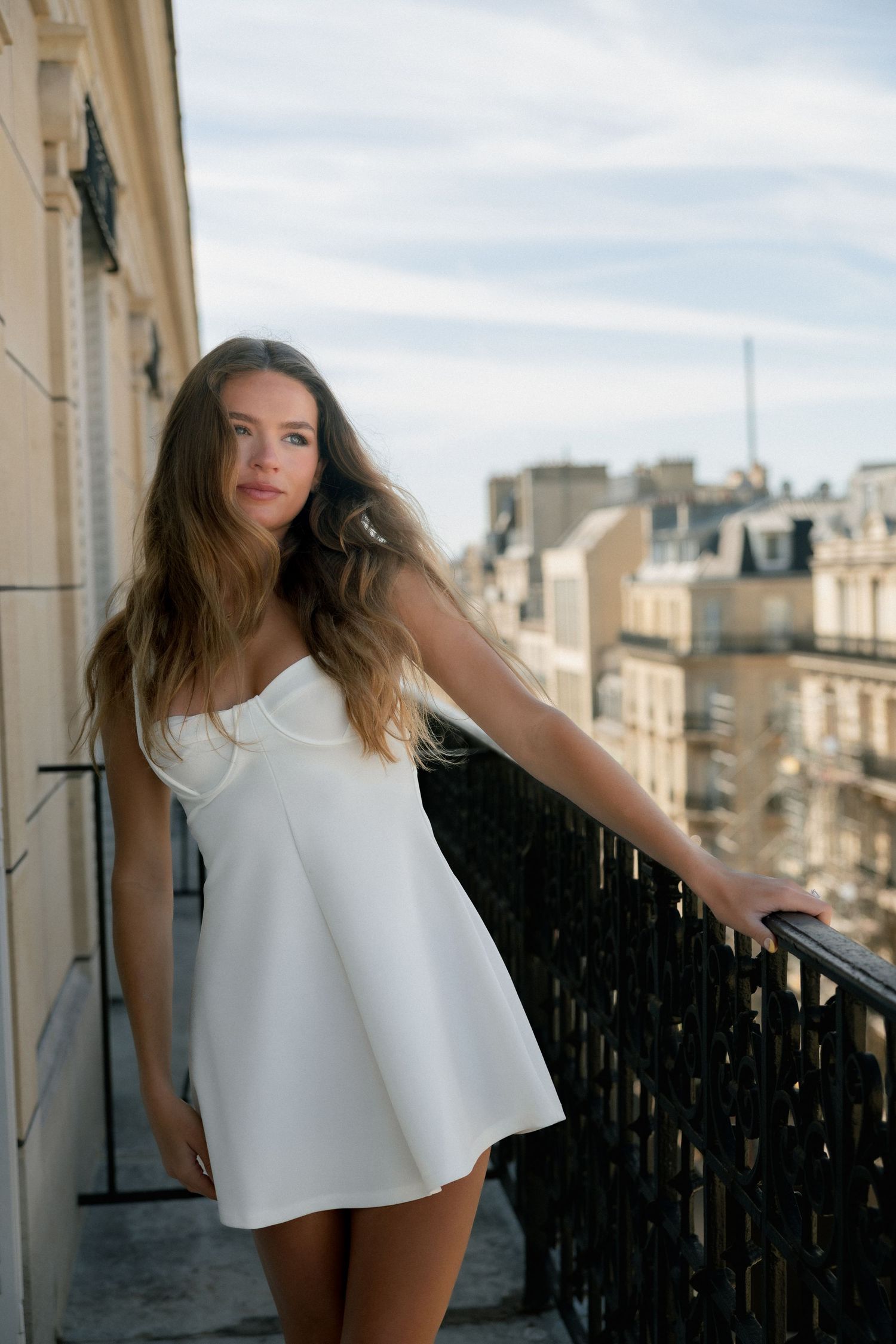 Young woman in white mini dress poses on Parisian balcony with city rooftops and Eiffel Tower in background.