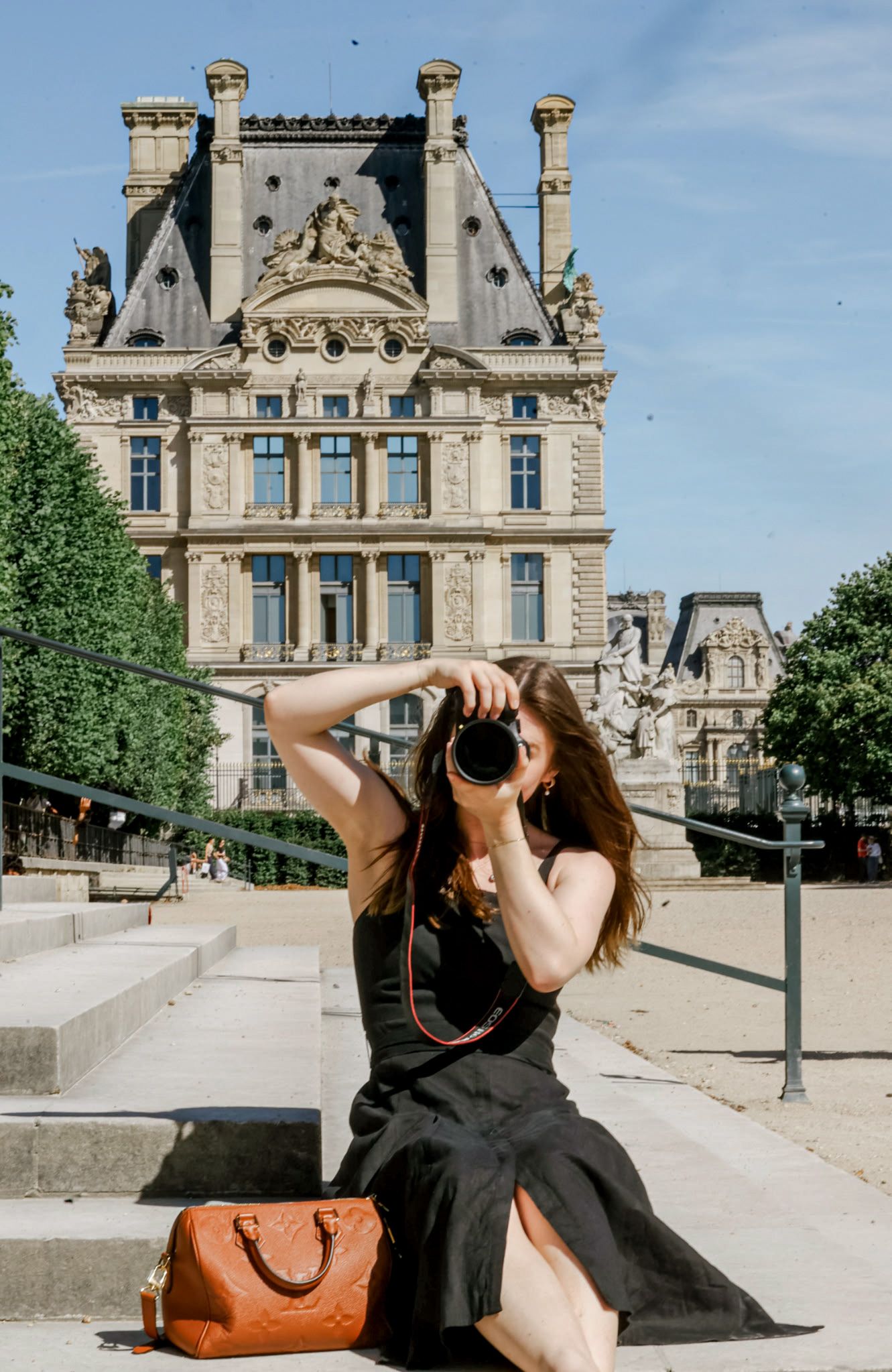 Woman in black dress photographing with camera on steps before ornate Parisian building with brown leather handbag nearby.