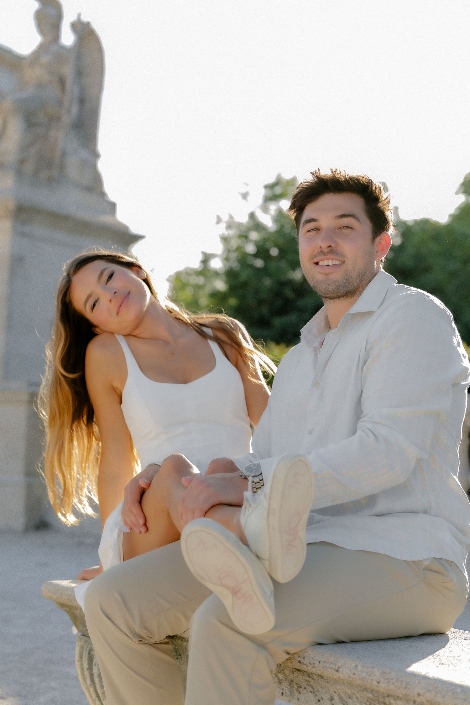 Couple sitting on stone ledge smiling near classical sculpture in golden sunlight.