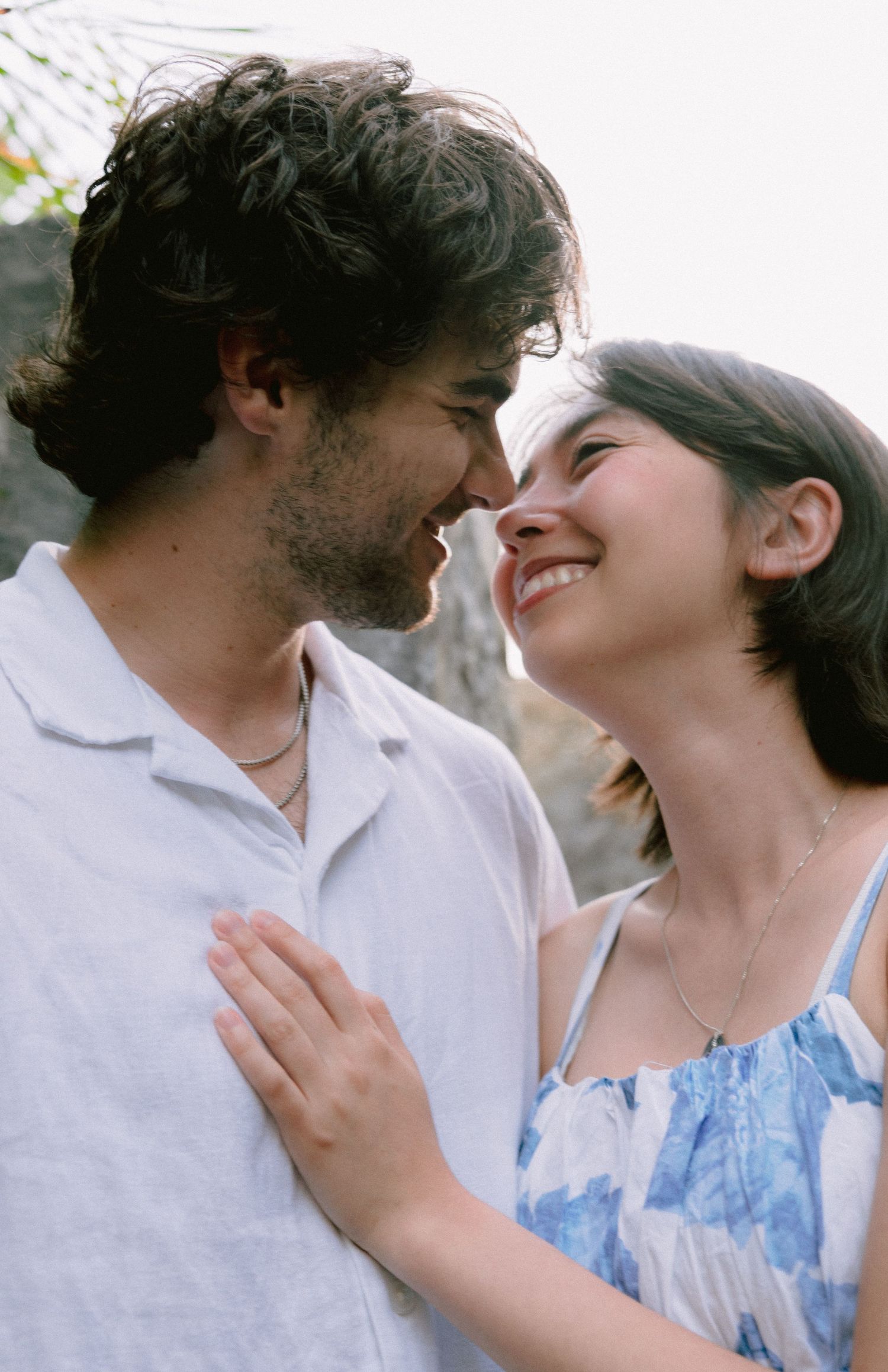 A smiling couple gazes lovingly at each other outdoors, the woman in a blue floral dress and the man in a white linen shirt.