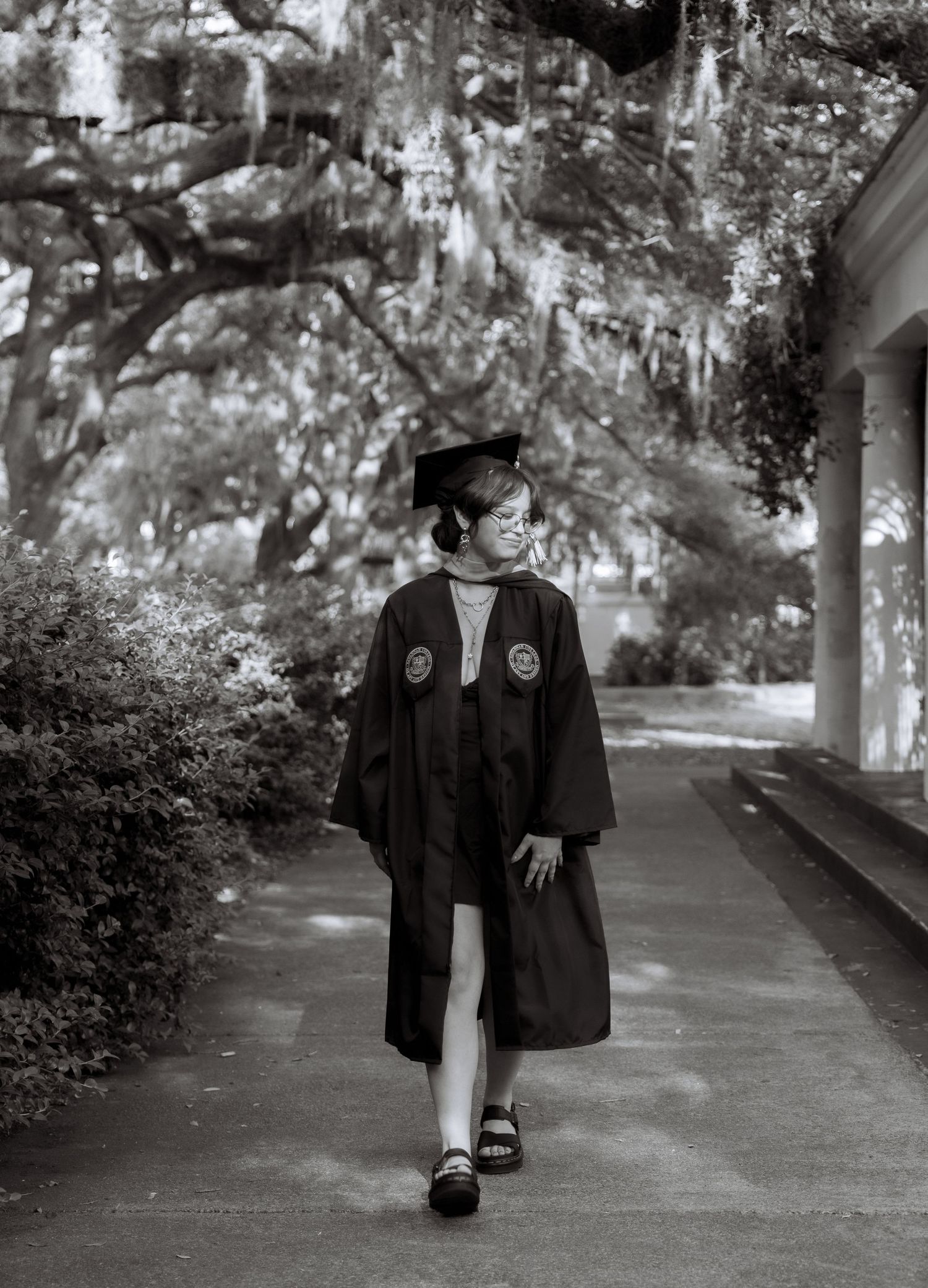 A graduate walks confidently in cap and gown along a tree-lined path in this black and white photo.