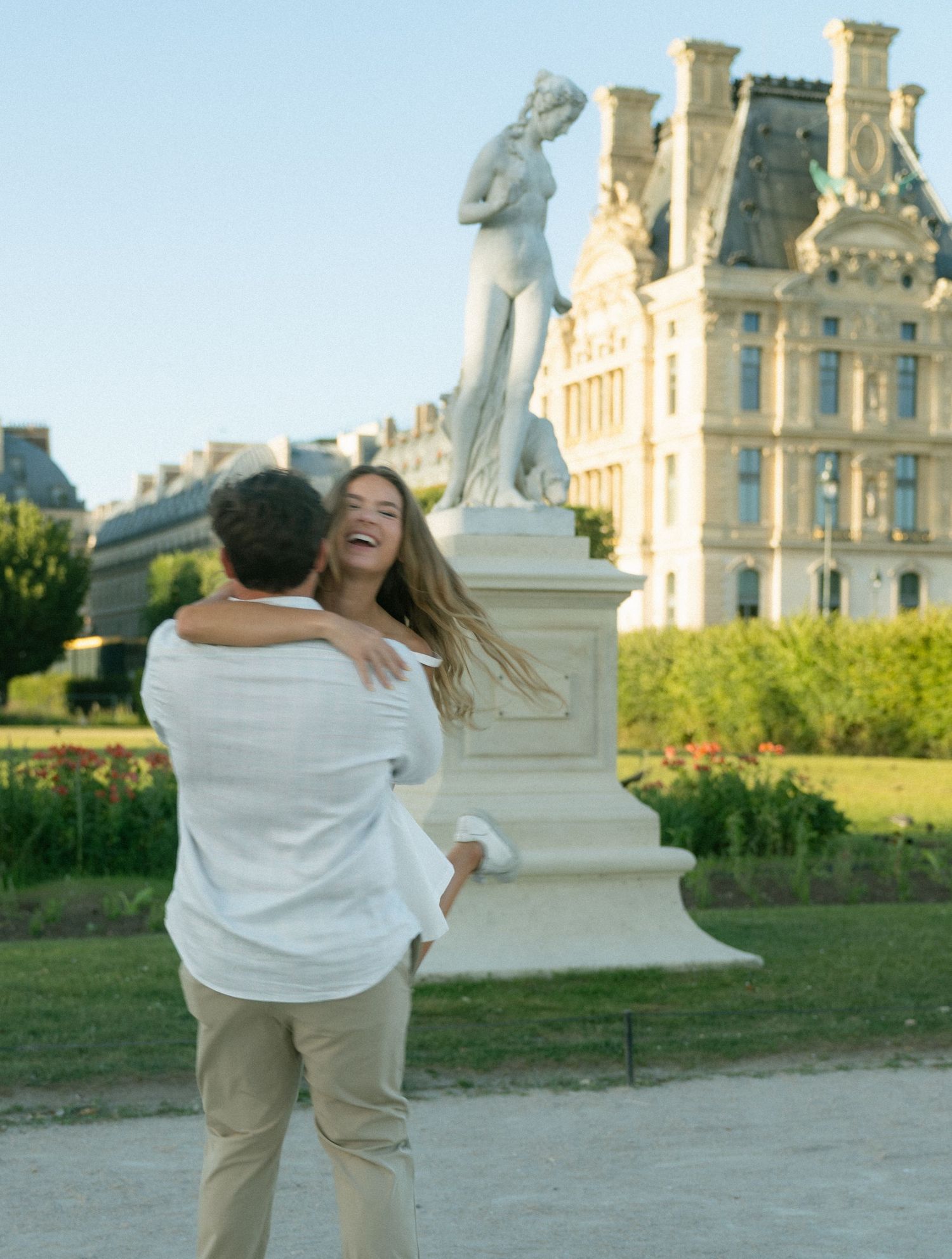 Couple laughing and embracing in Tuileries Garden with a white marble statue and Louvre Palace behind.