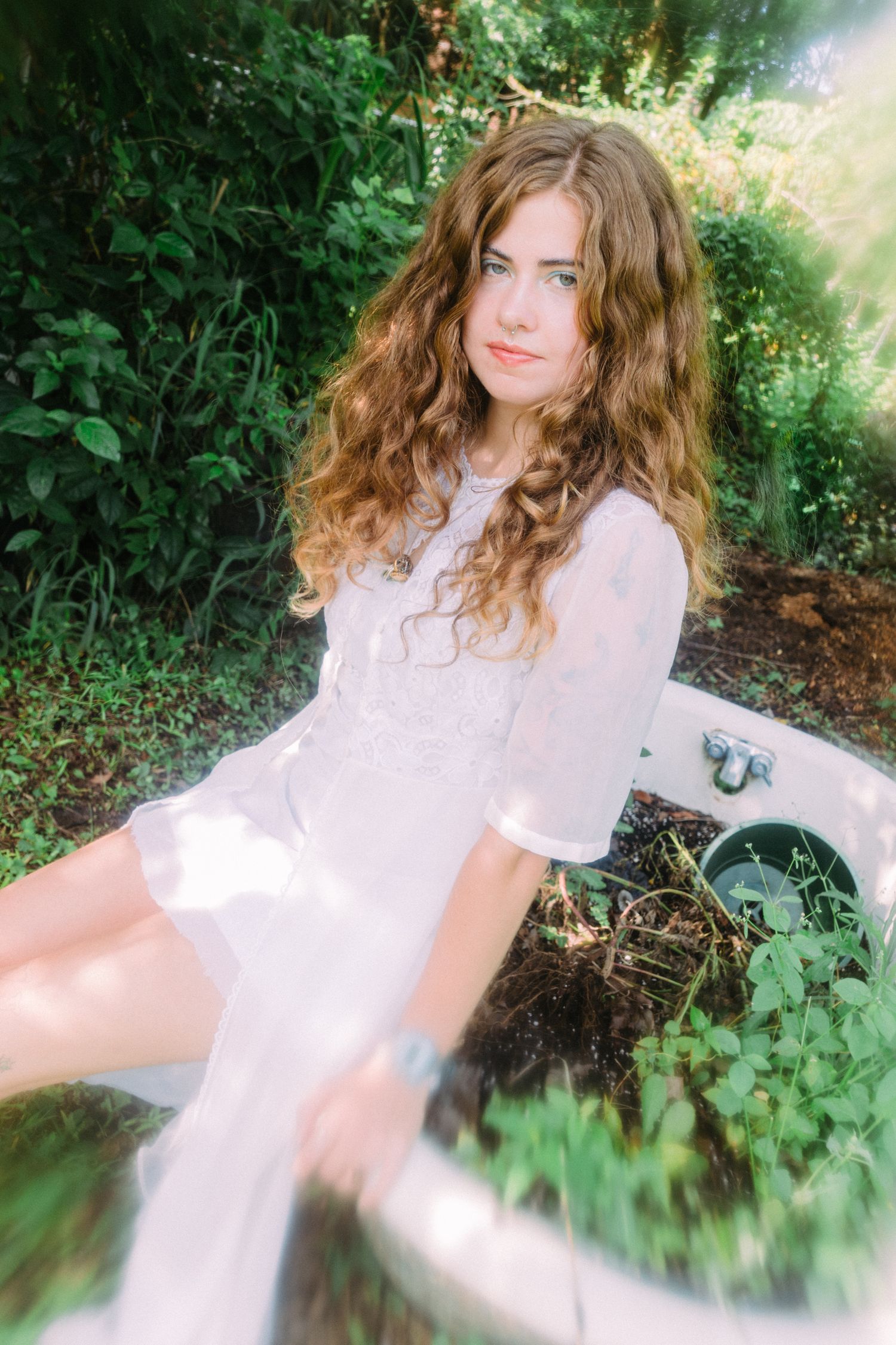 Young woman with curly hair in white lace dress sitting outdoors among lush green plants and foliage.