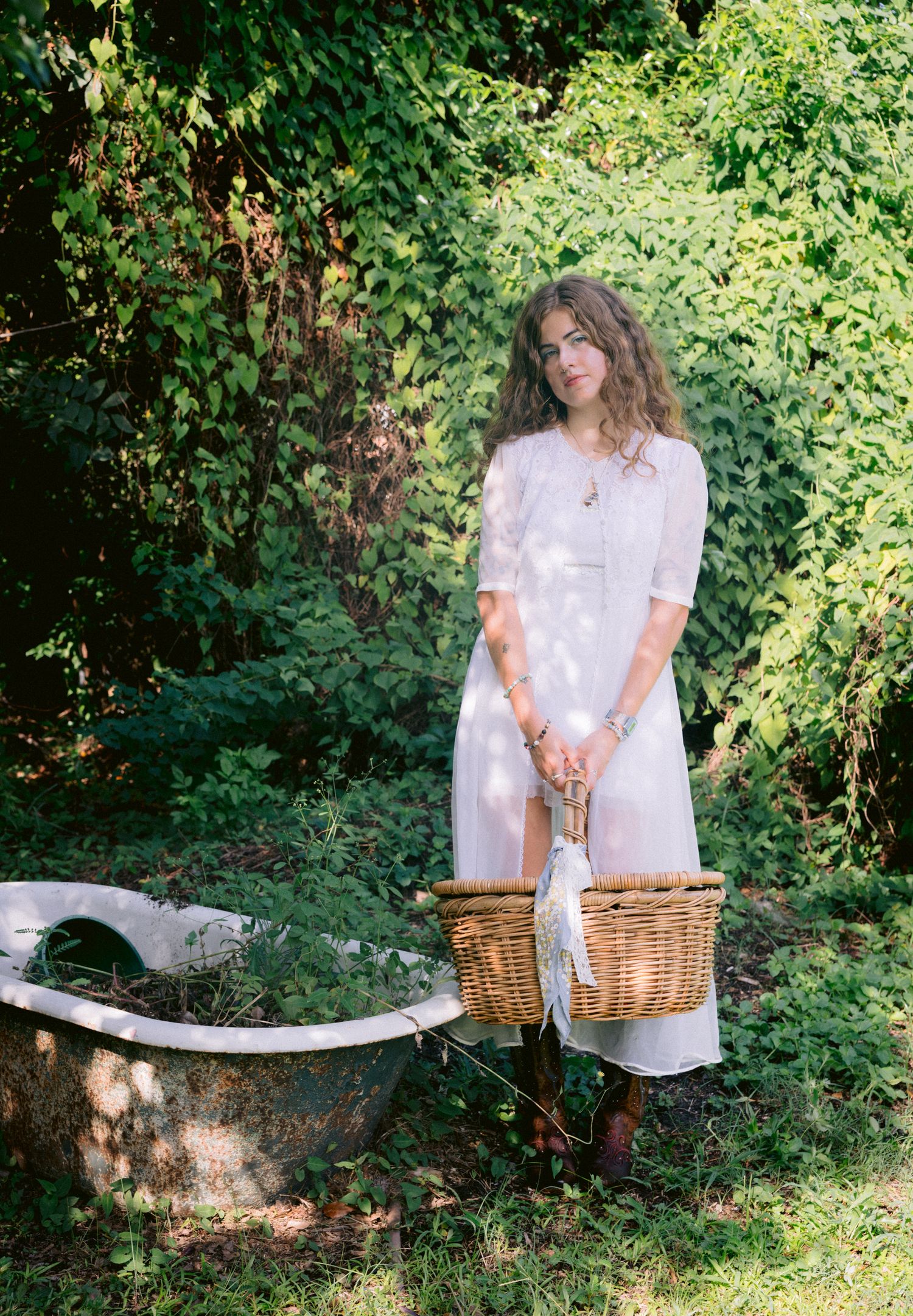 Woman in white dress holding wicker basket stands near vintage clawfoot bathtub surrounded by lush green foliage.