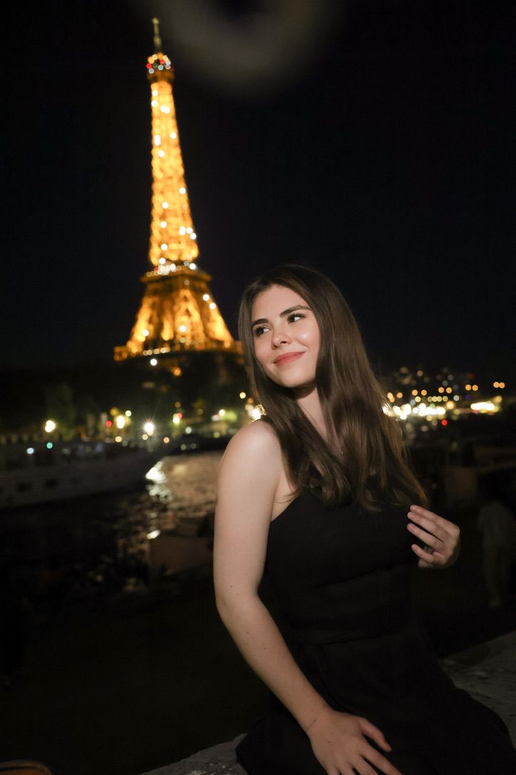Young woman in black dress smiling at night with illuminated Eiffel Tower glowing gold behind her in Paris.