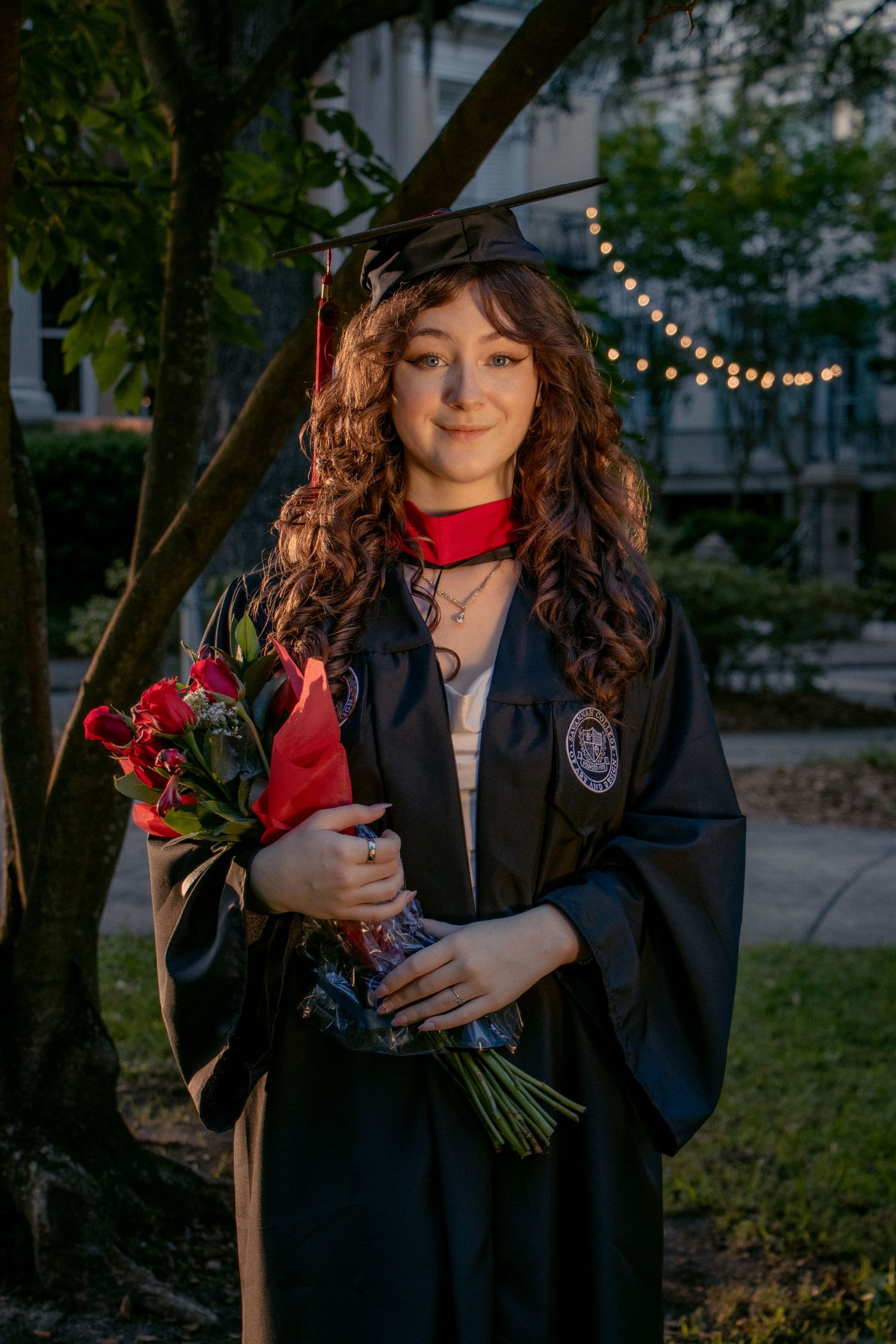 A curly-haired graduate in cap and gown holds red roses with string lights glowing behind her.