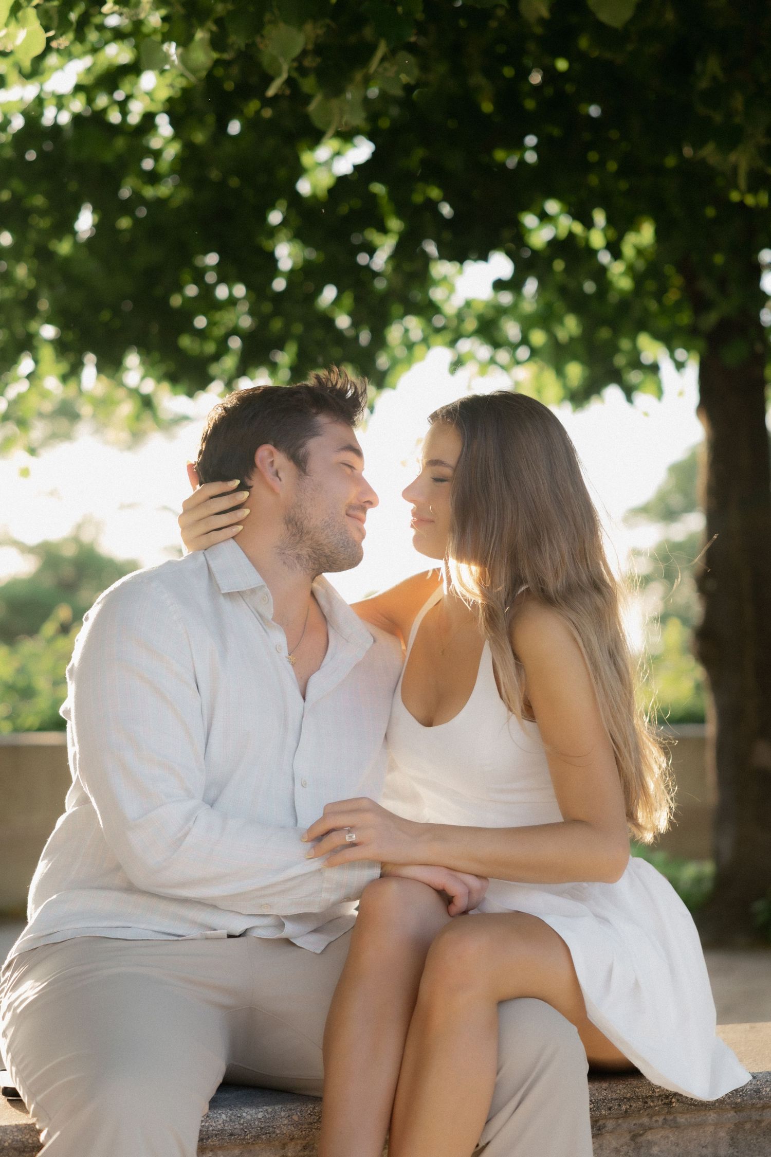 Smiling couple sitting close together under lush green trees in a sunny Parisian garden setting.