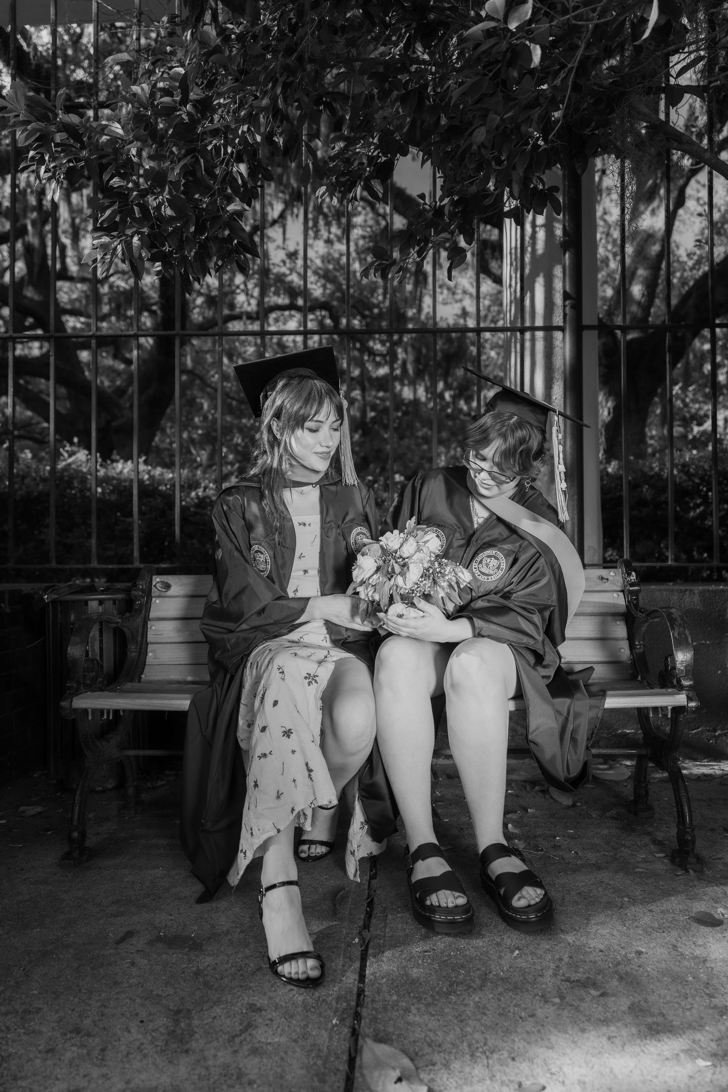 Two graduates in caps and gowns sit on a bench sharing a bouquet of flowers in black and white.