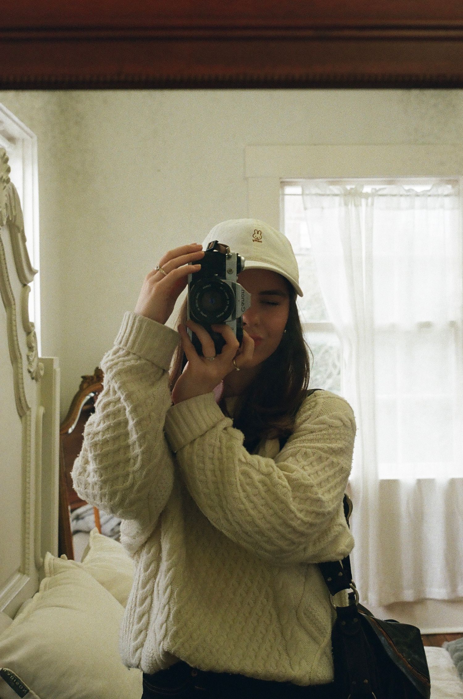 A young woman in a cream cable-knit sweater and white cap takes a mirror selfie with a film camera.