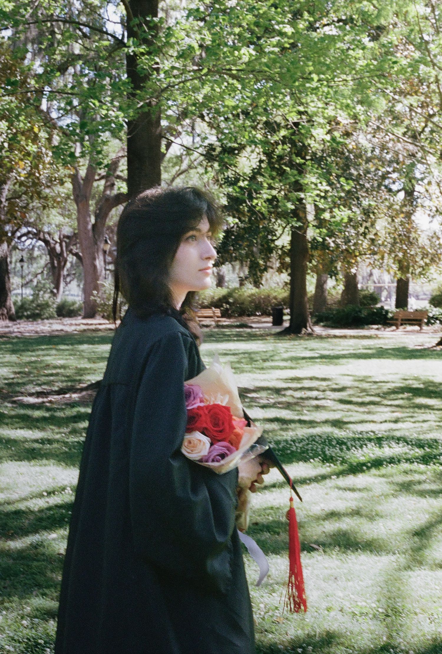 A graduate in a black gown holds a colorful bouquet while gazing into the distance in a sunny park.