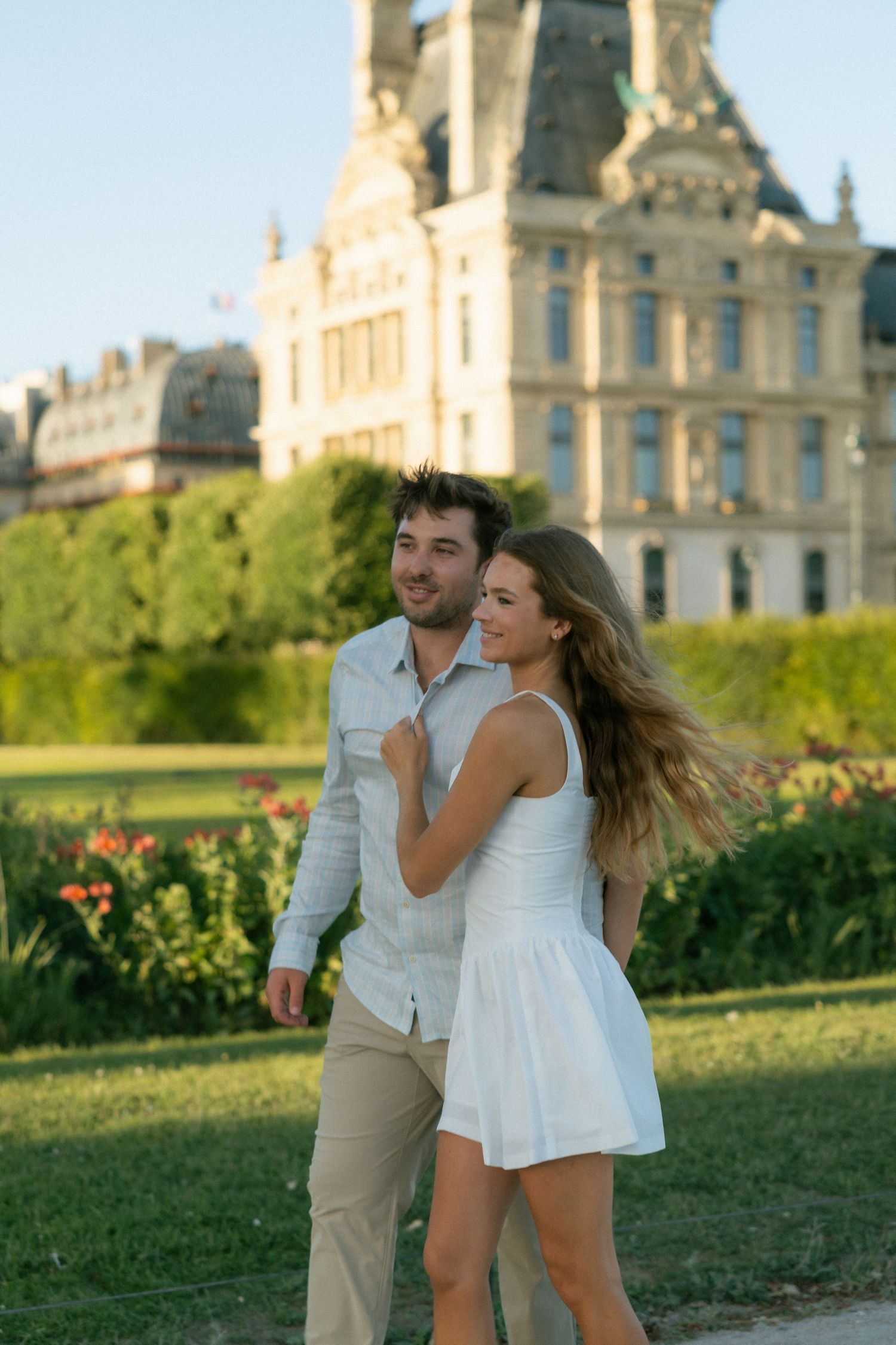 Happy couple walking together in Tuileries Garden with the grand Louvre Palace glowing at golden hour.