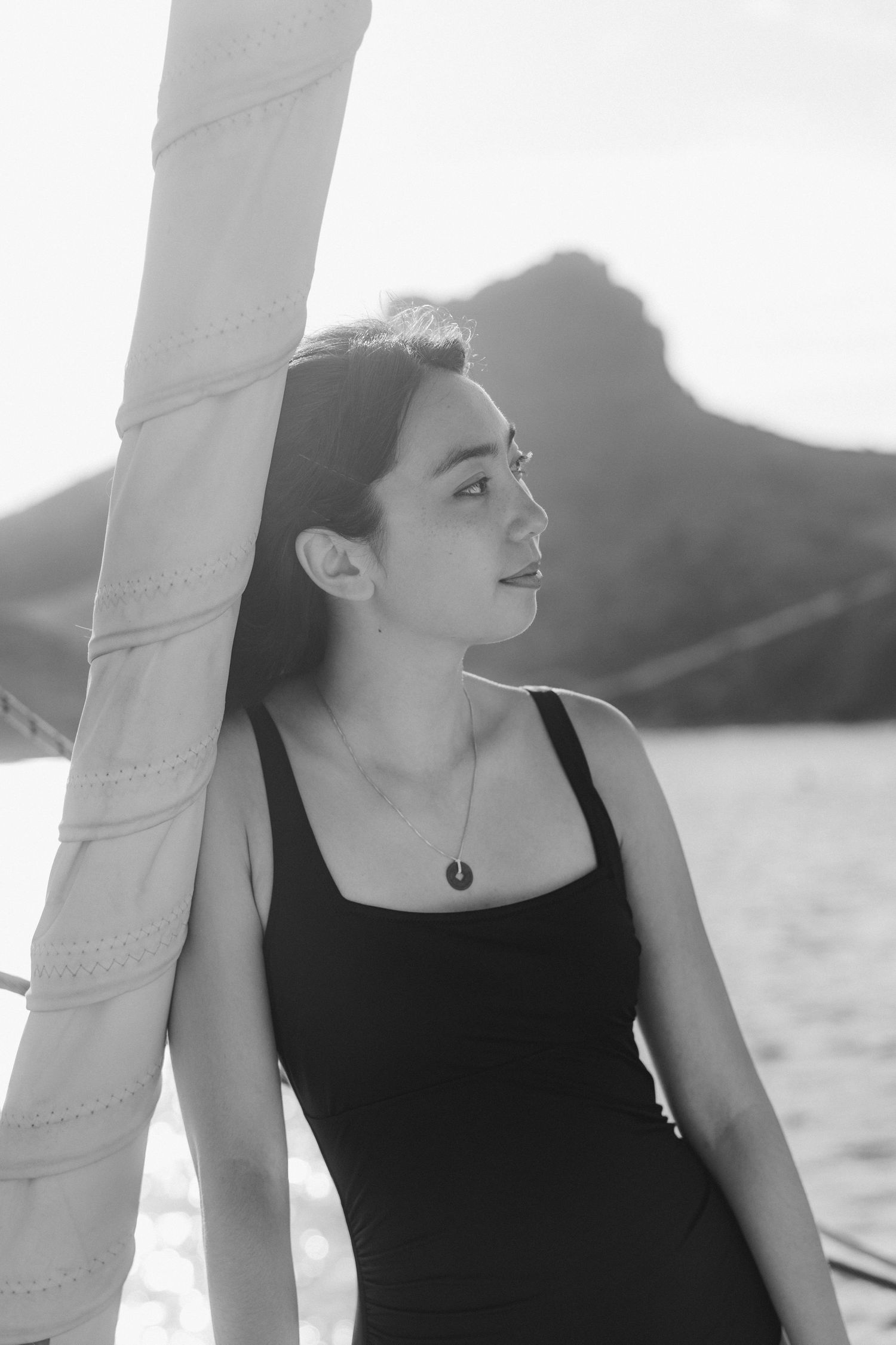 Black and white photo of a young woman in a swimsuit on a sailboat, gazing into the distance with mountains behind her.