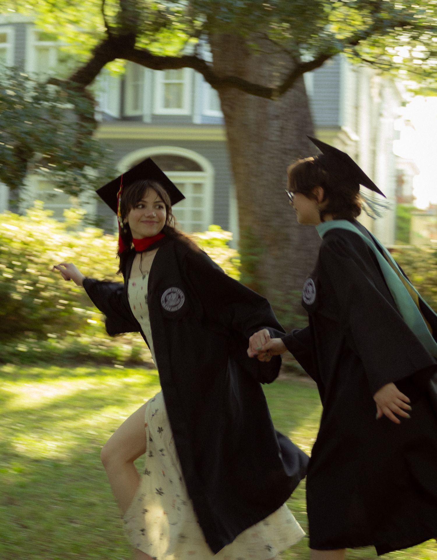 Two graduates in black caps and gowns run hand-in-hand across a sunlit green lawn together.