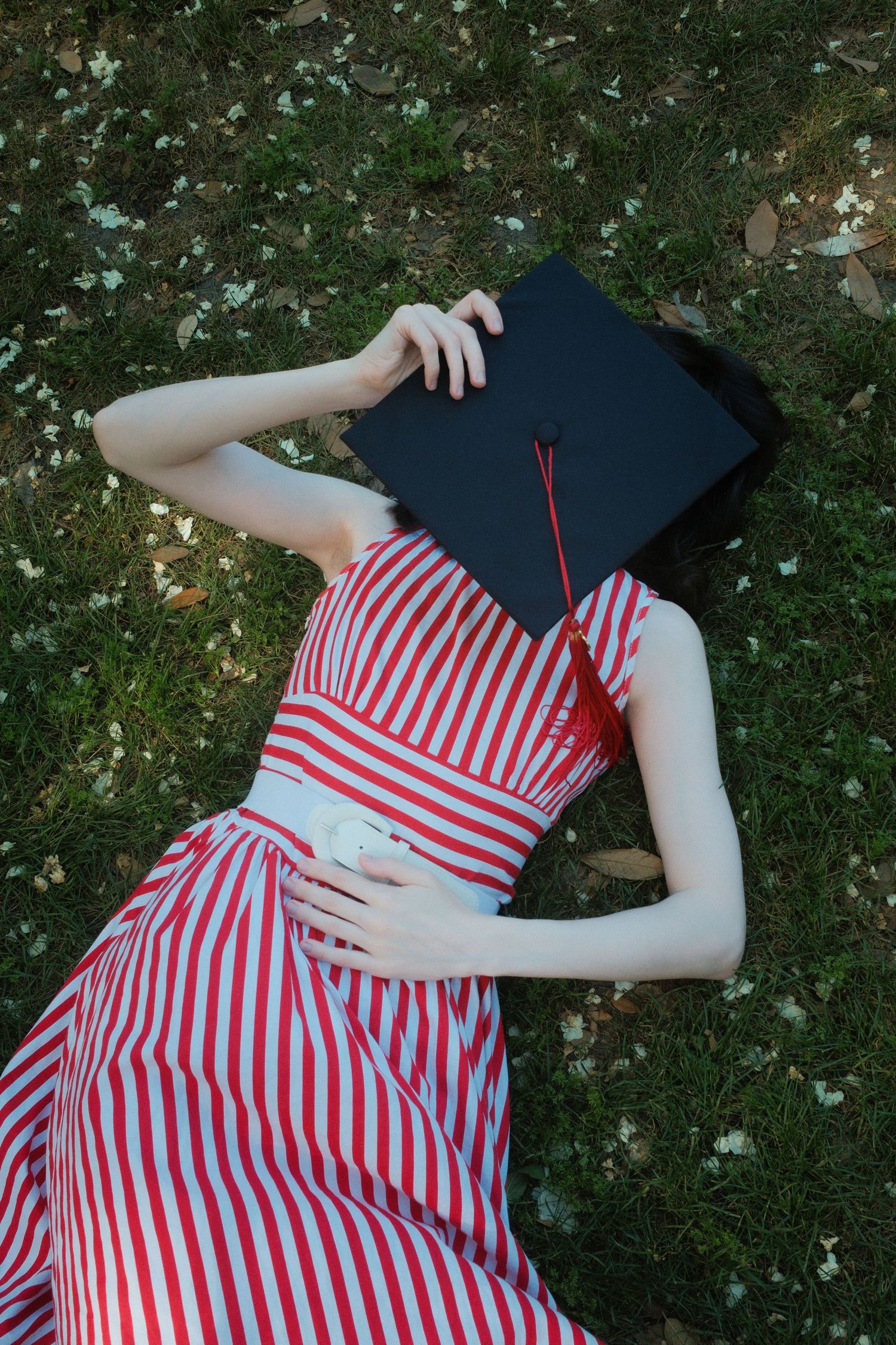 A graduate in a red striped dress lies on grass holding a black mortarboard cap over her face.