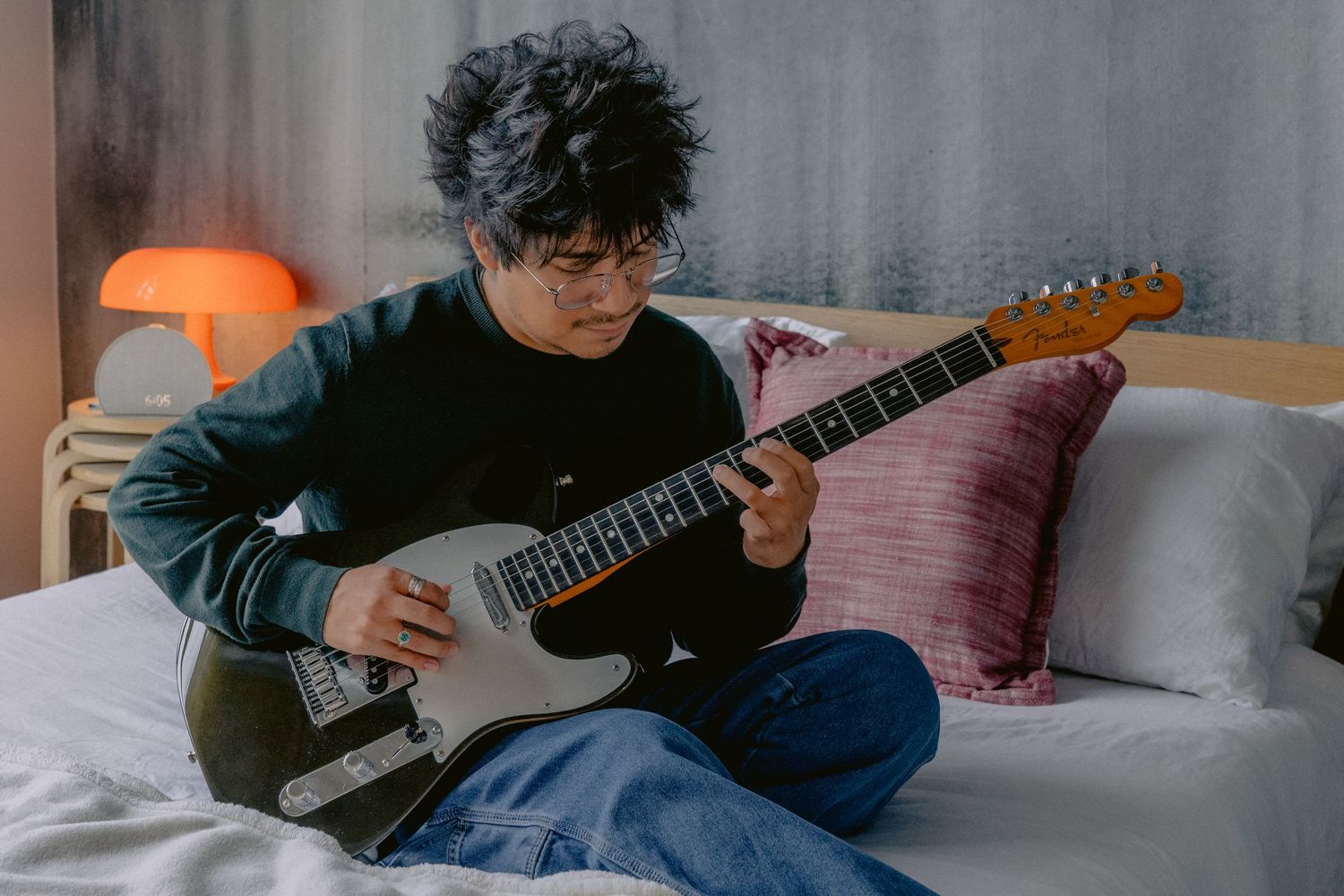 Young person with curly hair and glasses playing electric guitar while sitting on a bed with colorful pillows.