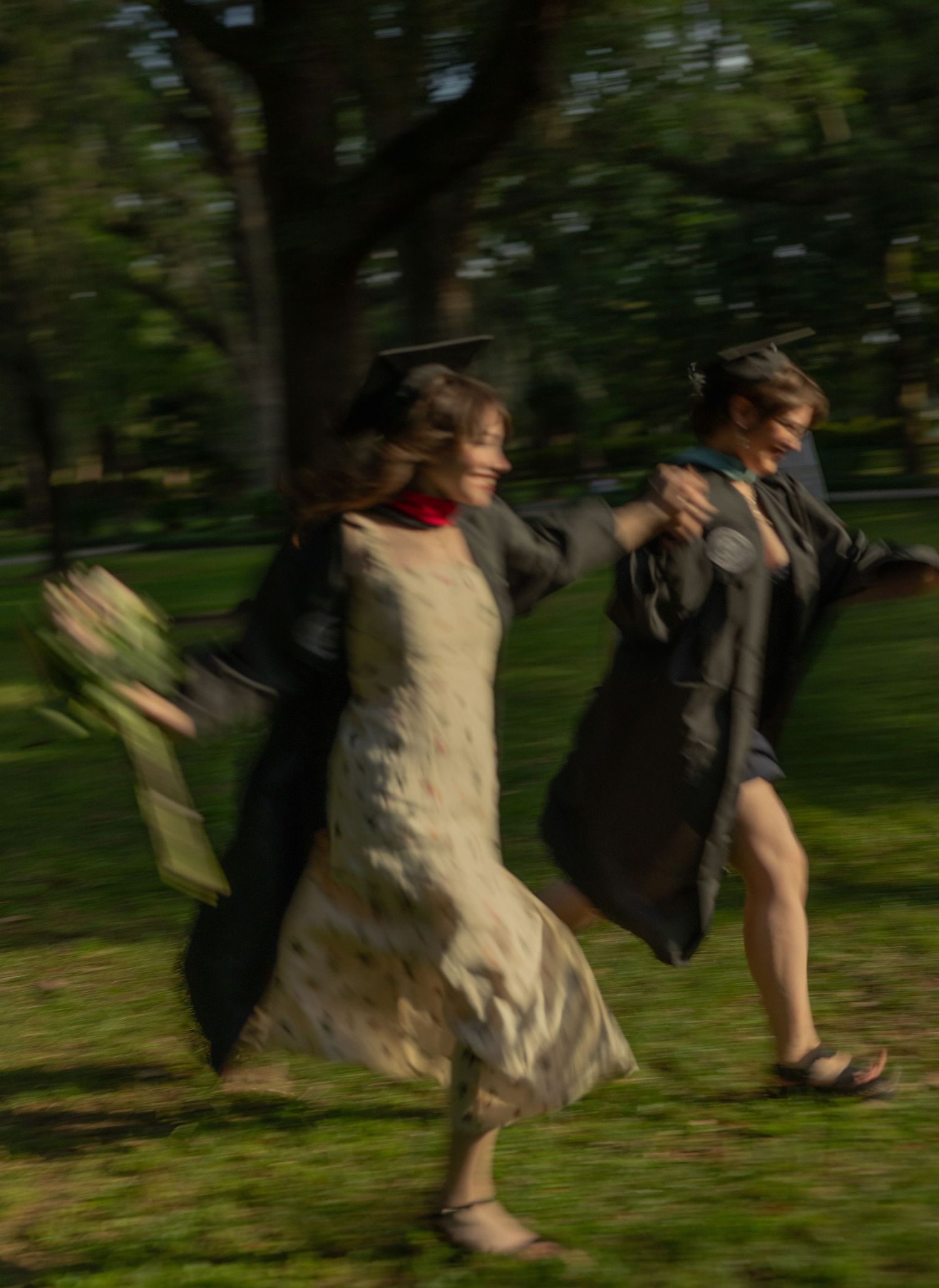 Two graduates in black robes and caps run joyfully through sunlit park in motion blur photo.