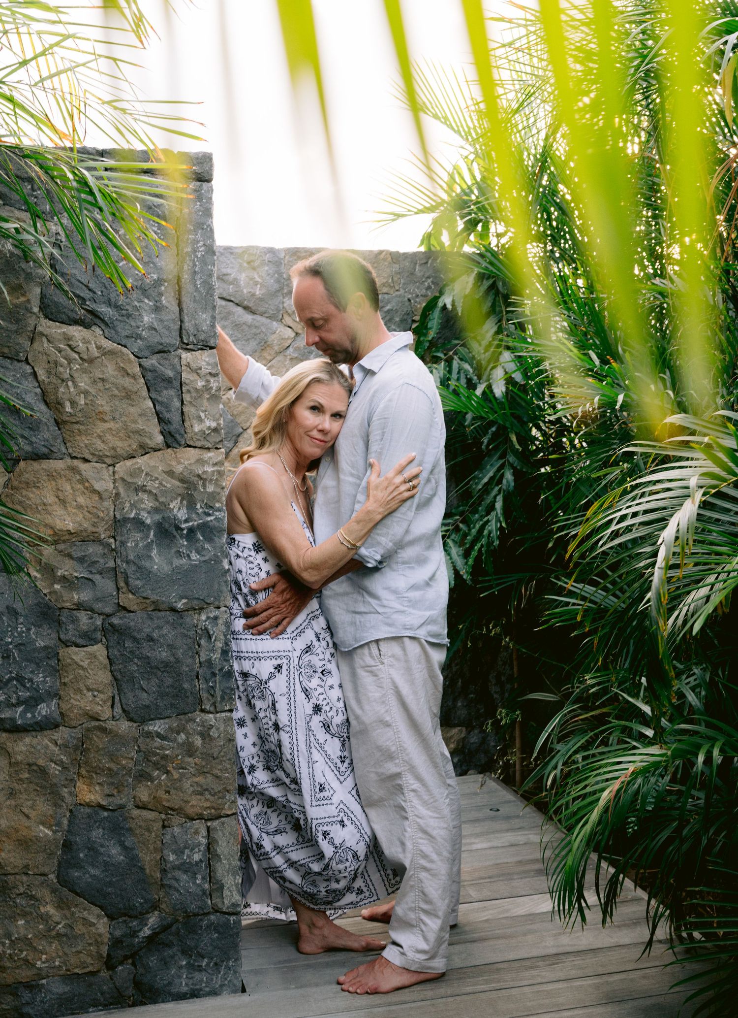 Couple embracing barefoot on wooden deck surrounded by tropical palms and stone wall in outdoor setting.