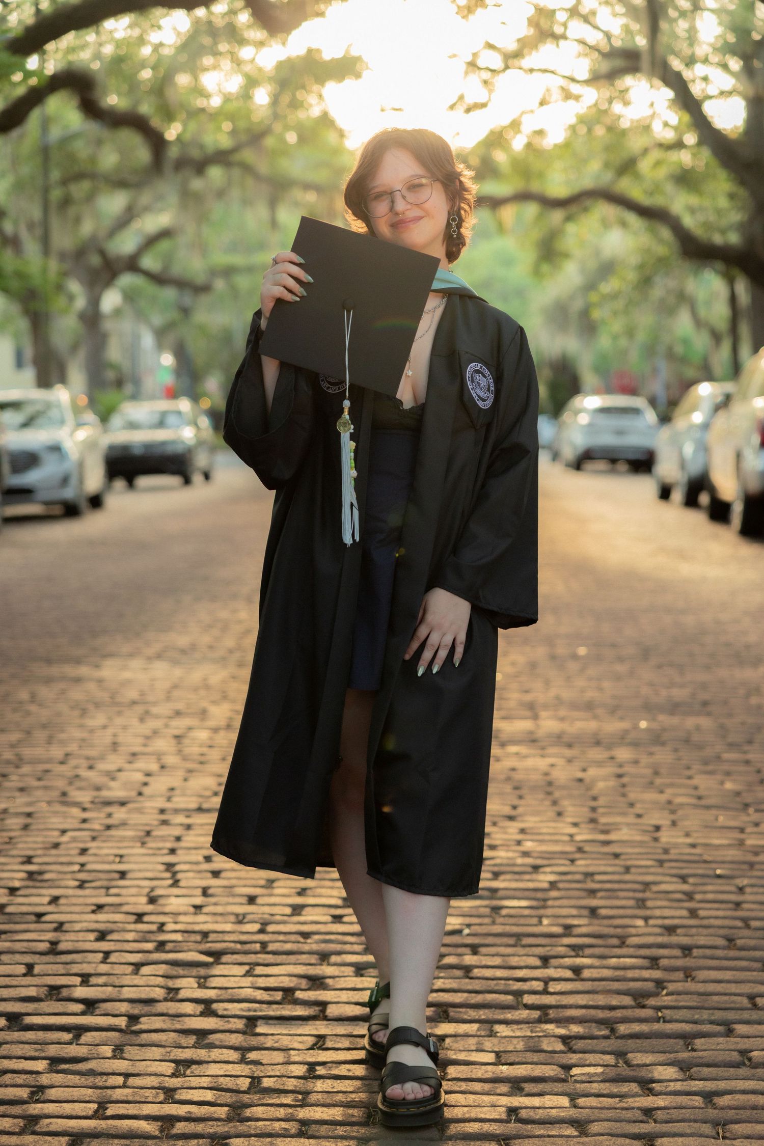 Graduate in black cap and gown holds diploma on tree-lined cobblestone street at golden hour.