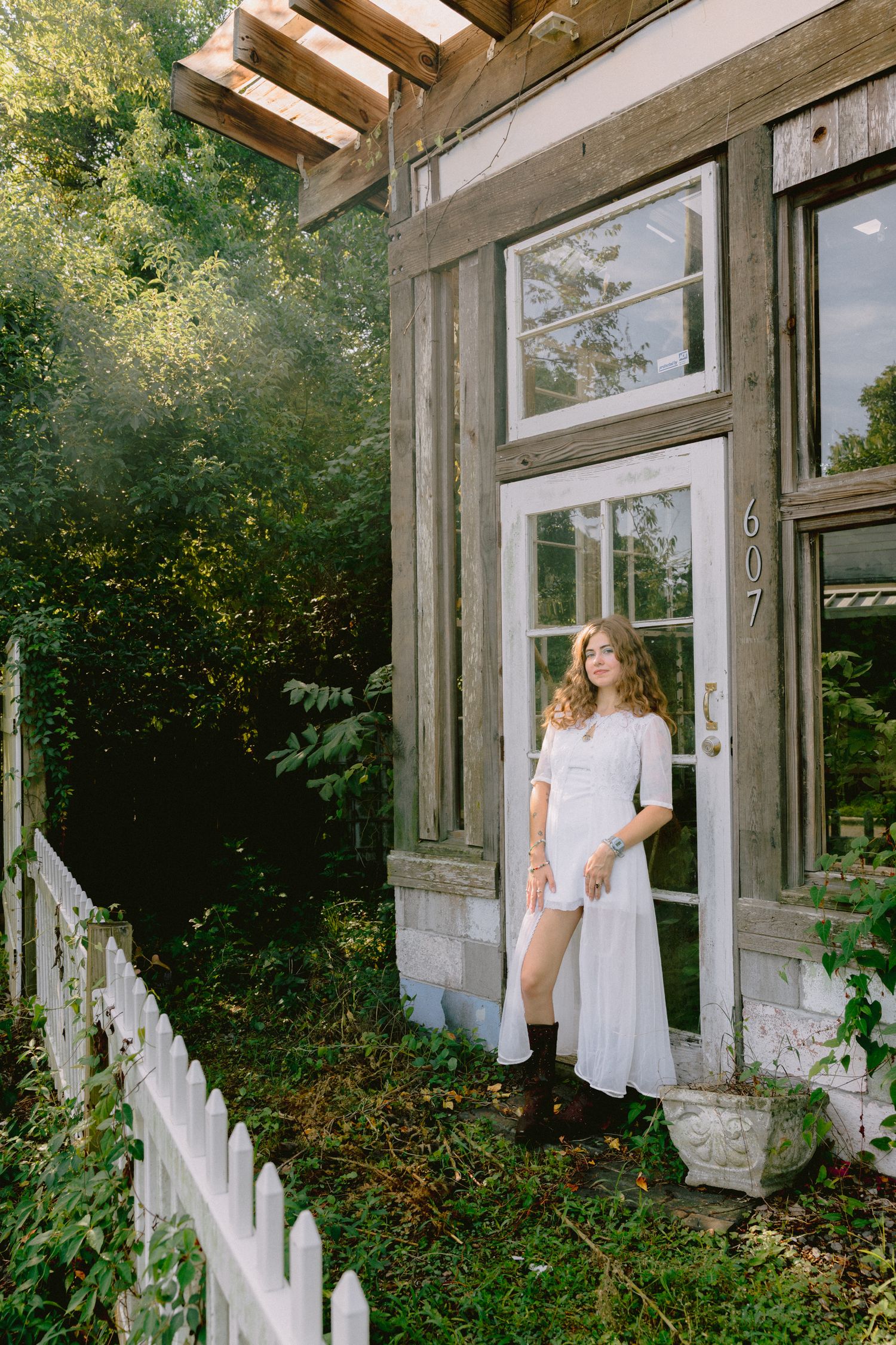 Young woman in white dress and boots stands at door 607 of rustic wooden building surrounded by lush greenery.