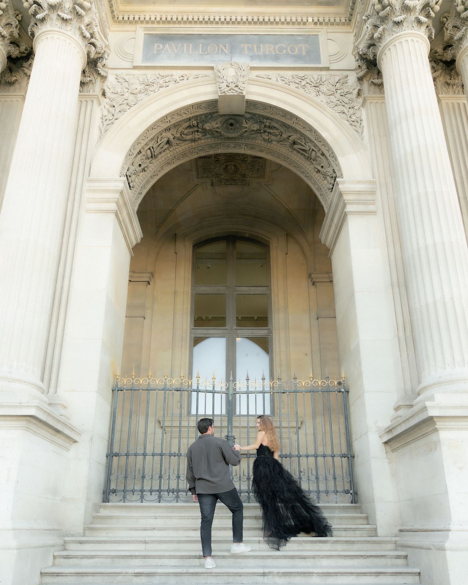 Elegant couple climbing ornate stone steps of the Petit Palais in Paris, woman in black gown.