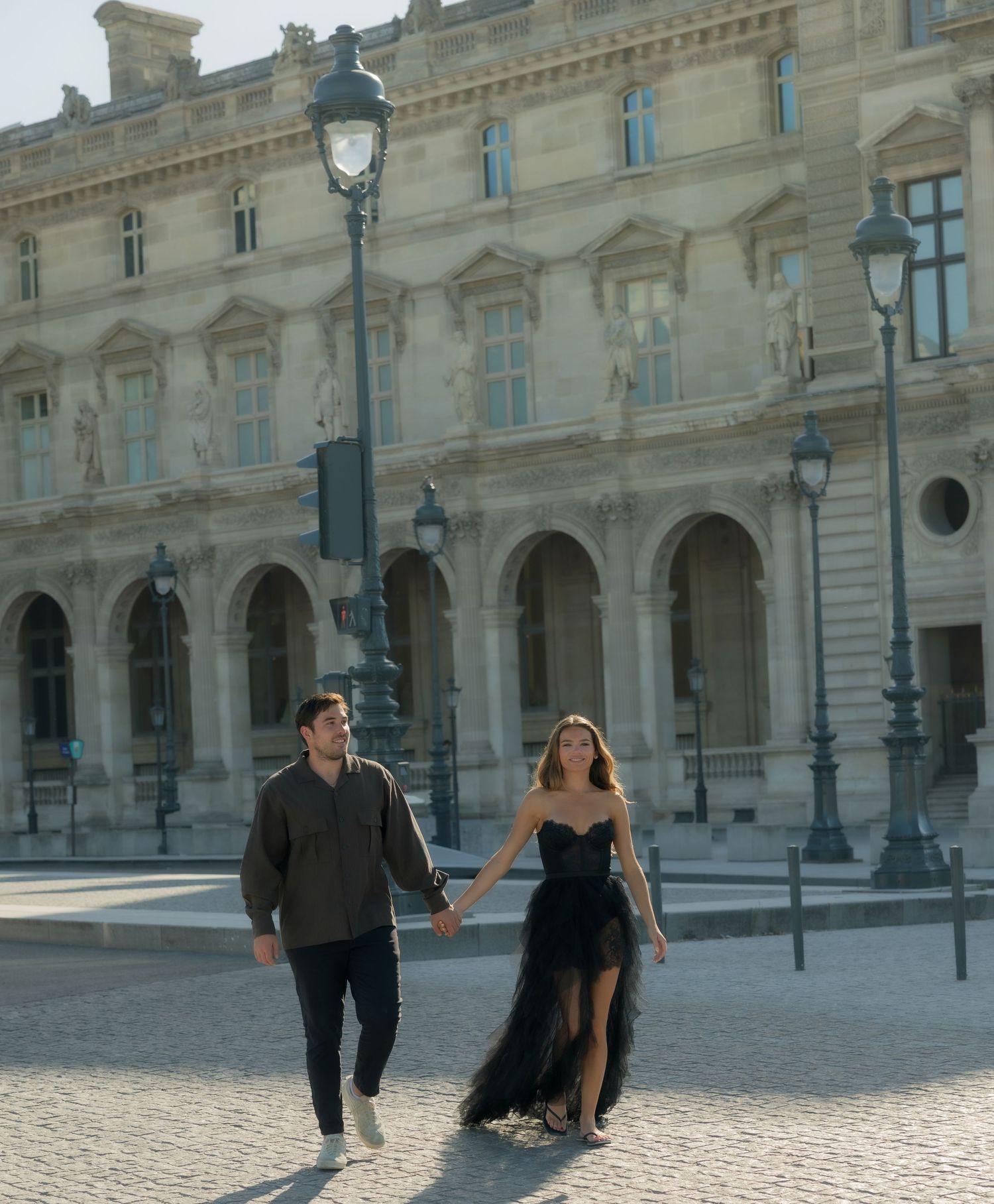 Couple holding hands walking along cobblestone path in front of the Louvre Museum in Paris.