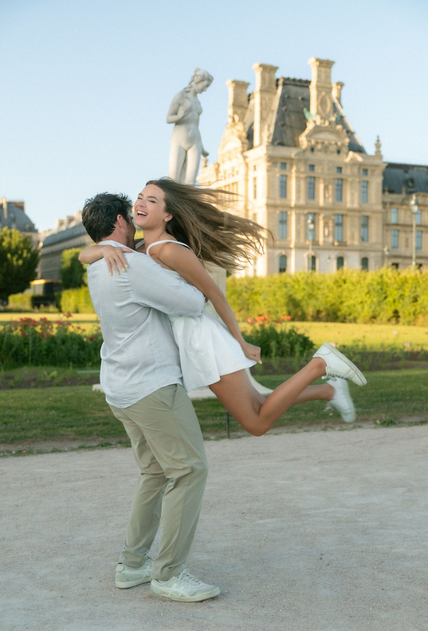 Man lifting smiling woman in white dress at Tuileries Garden with Louvre Palace in sunny background.