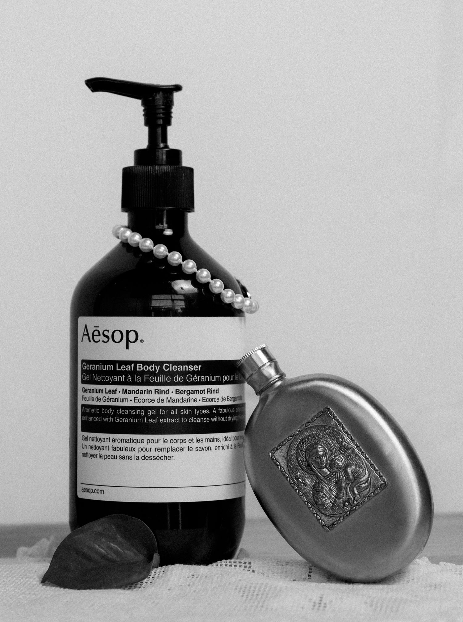 Black and white photo of Aesop Geranium Leaf Body Cleanser pump bottle with pearl necklace and vintage metal flask.