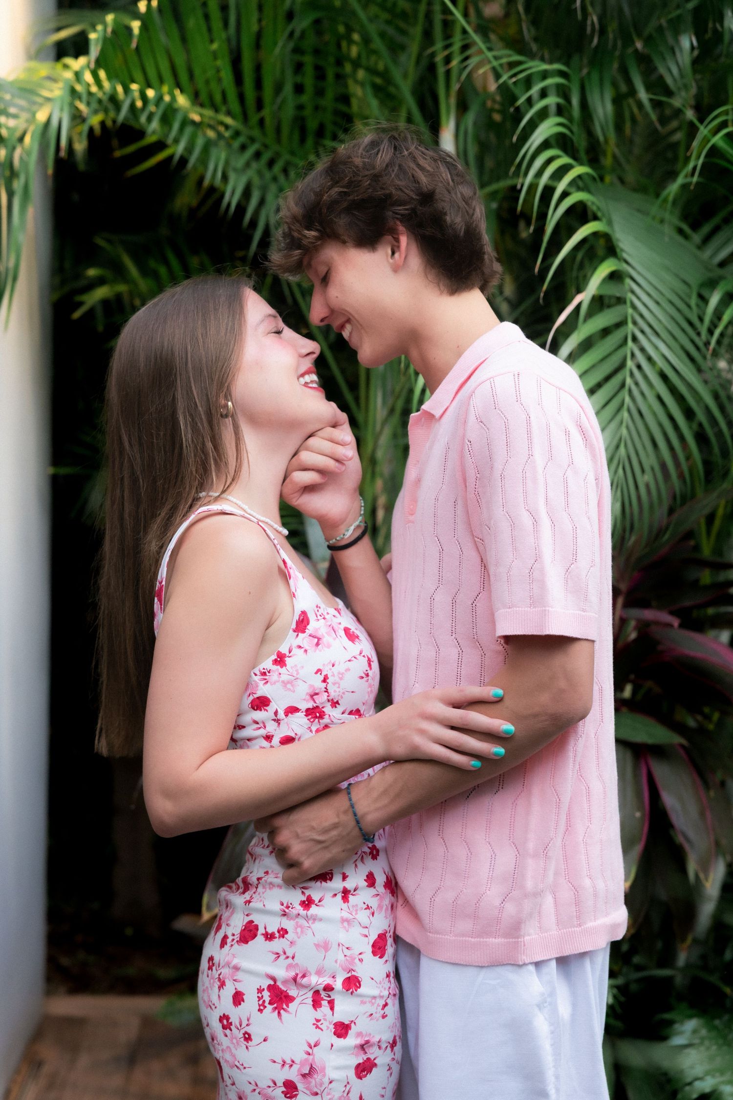 Young couple in pink and floral outfits sharing a romantic moment outdoors surrounded by tropical greenery.