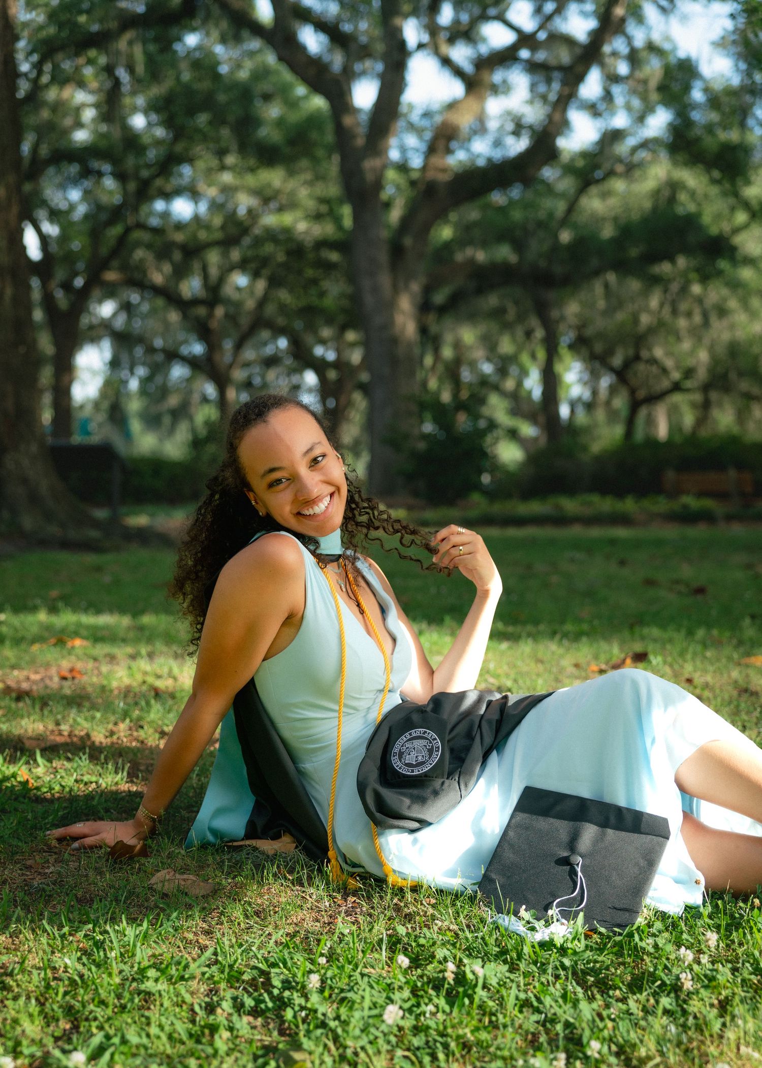 Smiling female graduate in light blue dress sits on grass in park with graduation gown.