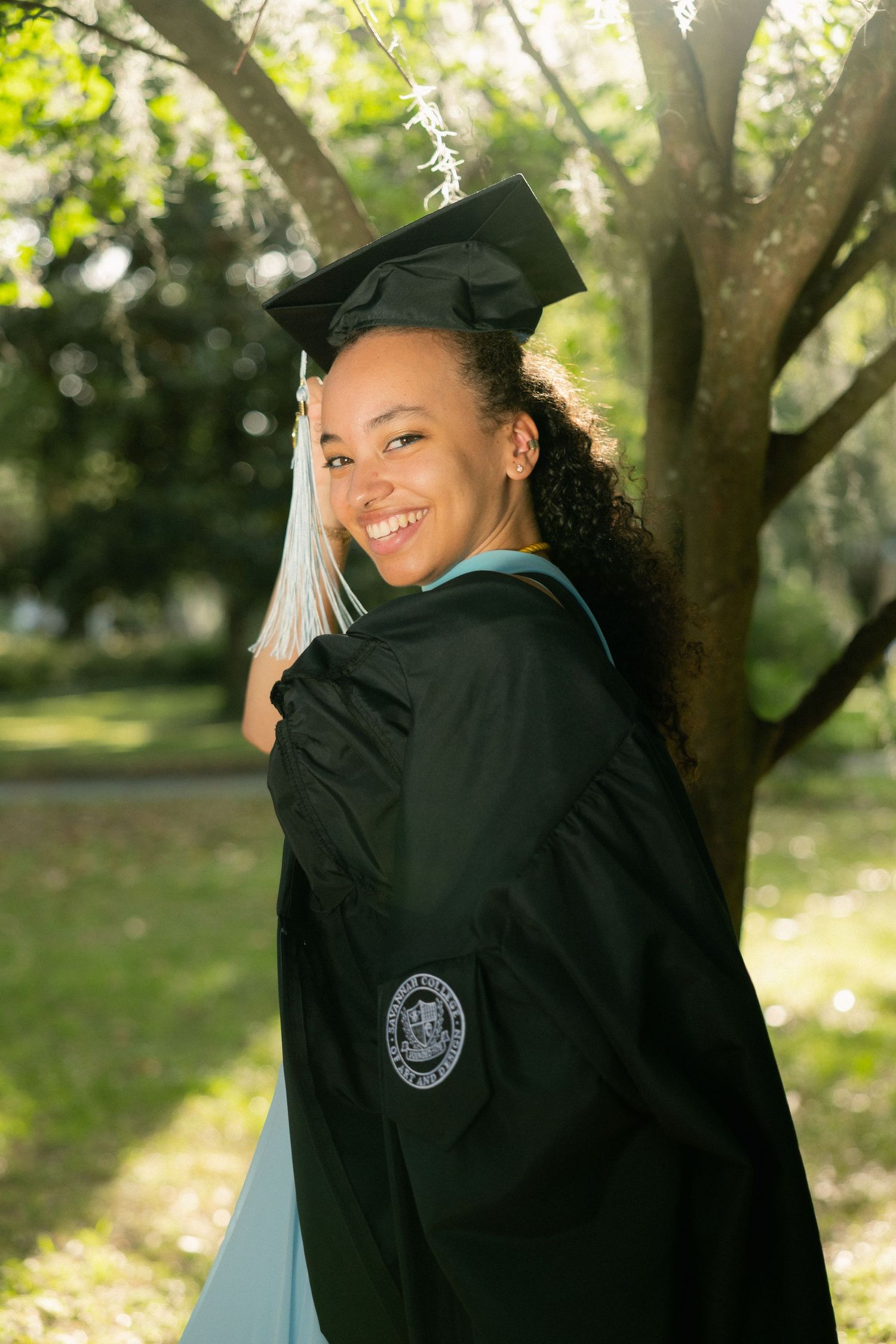 A beaming graduate glances back over her shoulder while wearing her black cap and gown outdoors.