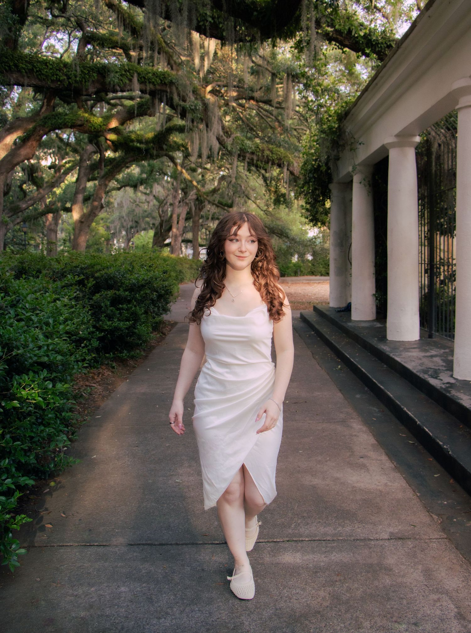 A young woman in a white wrap dress walks along a columned pathway under moss-draped oak trees.