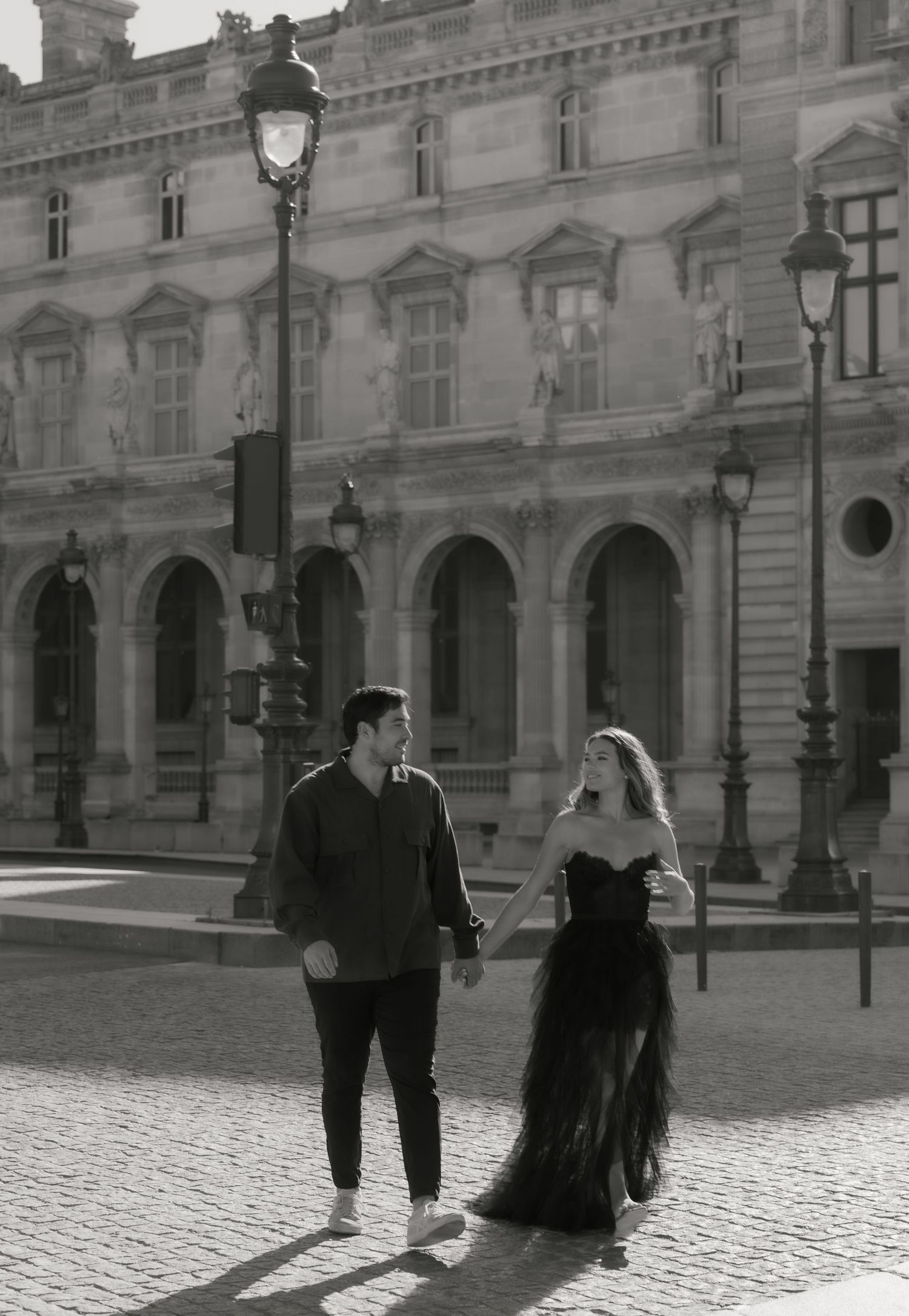 A couple holds hands walking past the grand Louvre museum in Paris in a black and white photo.
