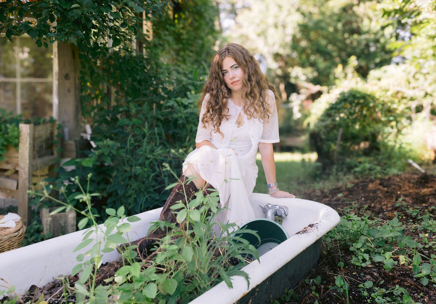 Woman in white outfit sitting in a vintage bathtub used as a garden planter surrounded by lush greenery.
