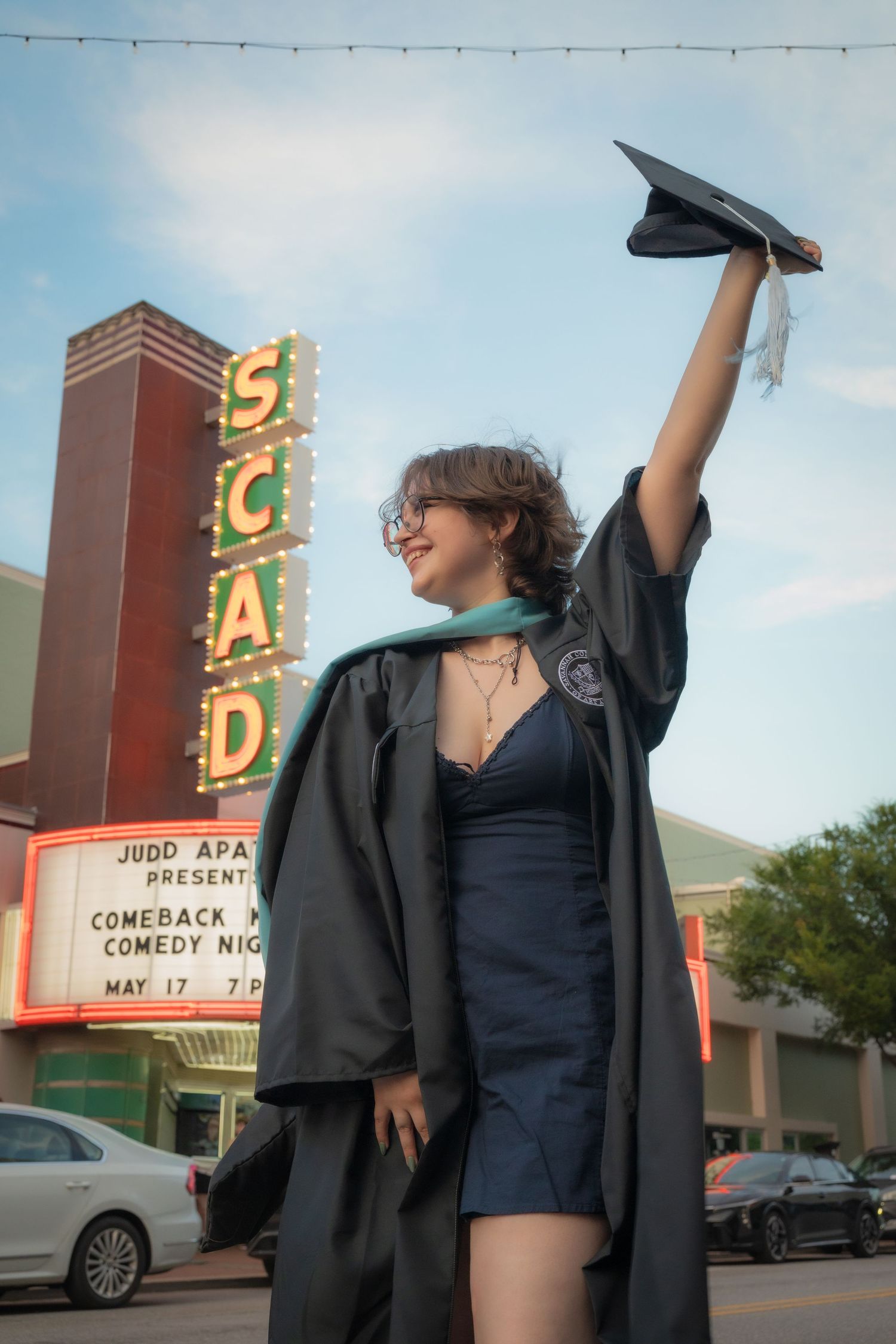 Female graduate in black robe raises cap triumphantly in front of illuminated SCAD theater marquee sign.