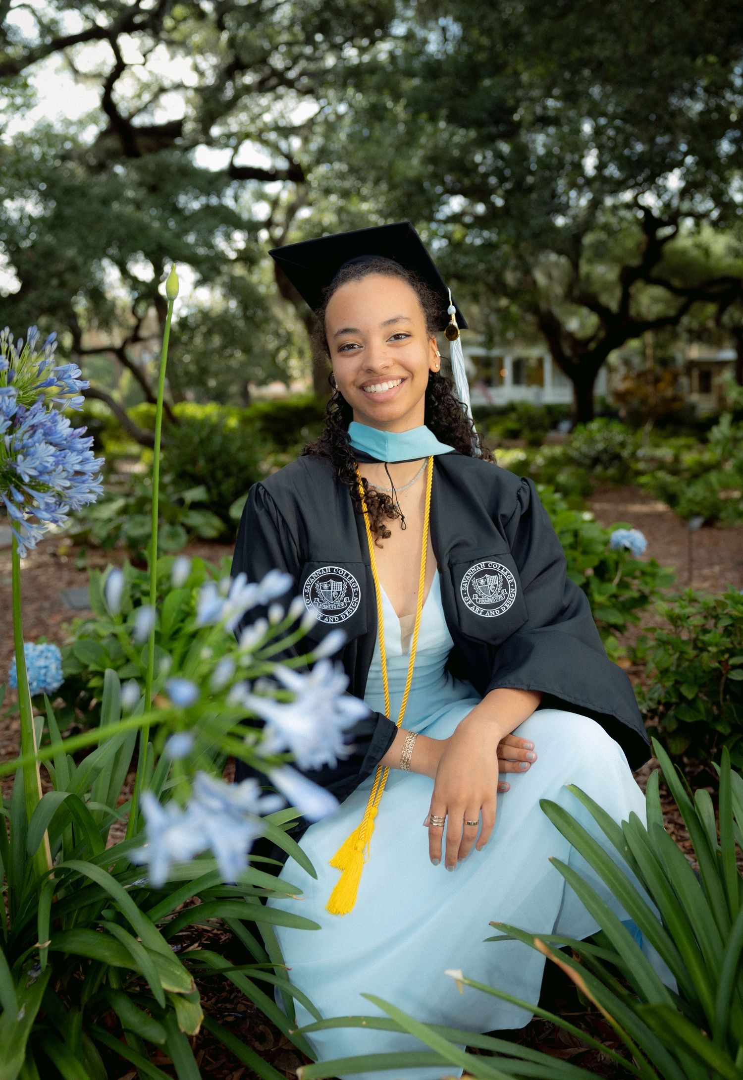 A smiling graduate in cap and gown with gold honor cord sits among blue agapanthus flowers.