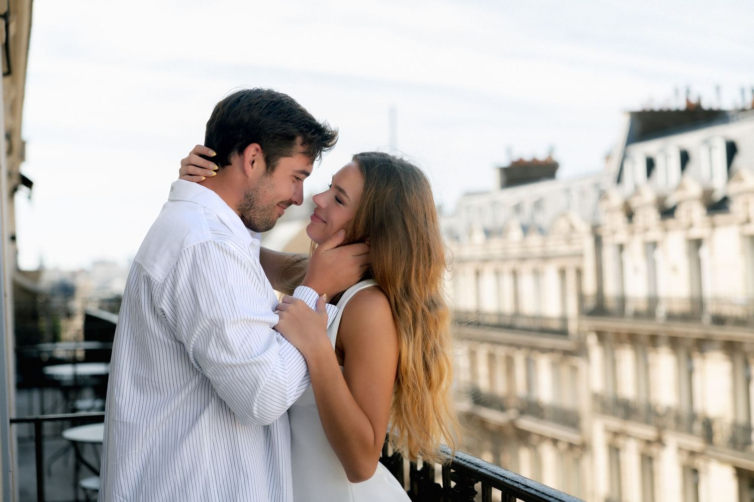 Romantic couple embracing on a Parisian balcony with classic Haussmann buildings and Eiffel Tower in background.