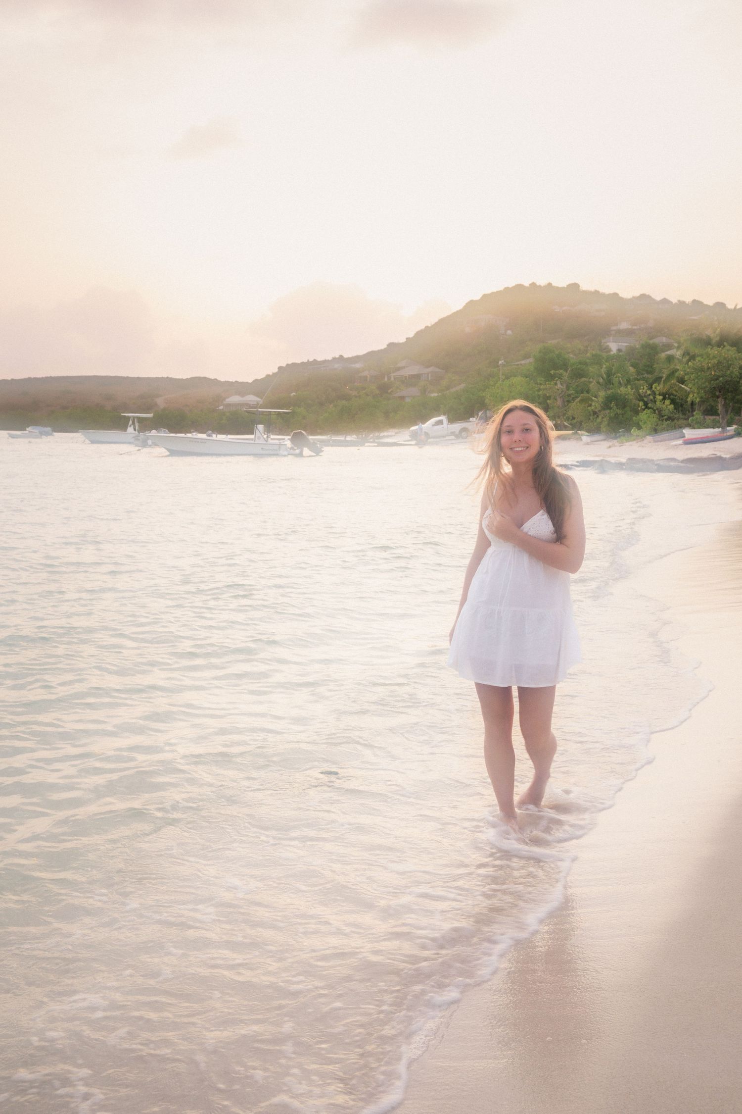 Young woman in white dress walking on tropical beach at sunset with boats and green hills in background.