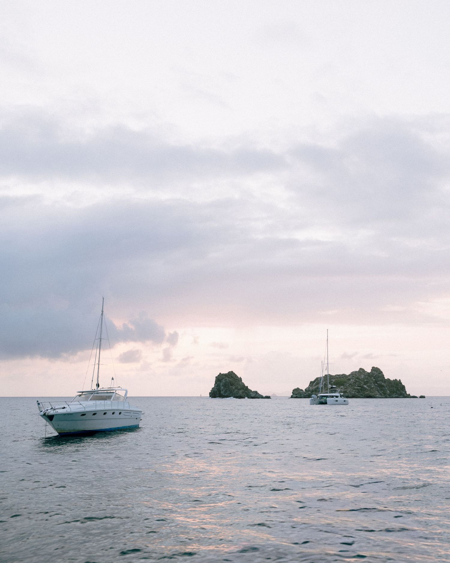 Two white sailboats anchored near rocky islands at sunset with soft pastel clouds over calm ocean waters.