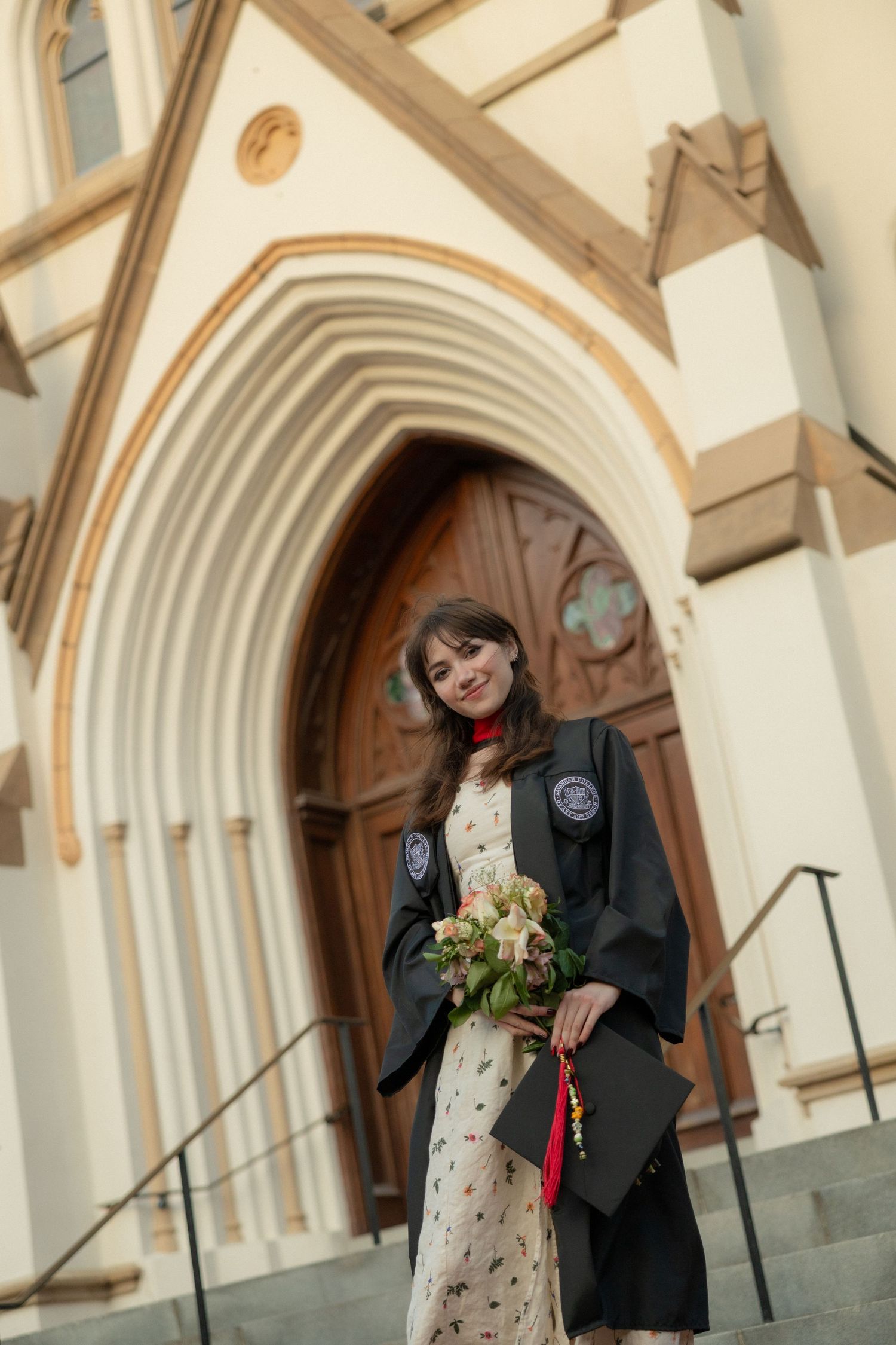 Female graduate in black robe holds flowers bouquet outside ornate church archway entrance.