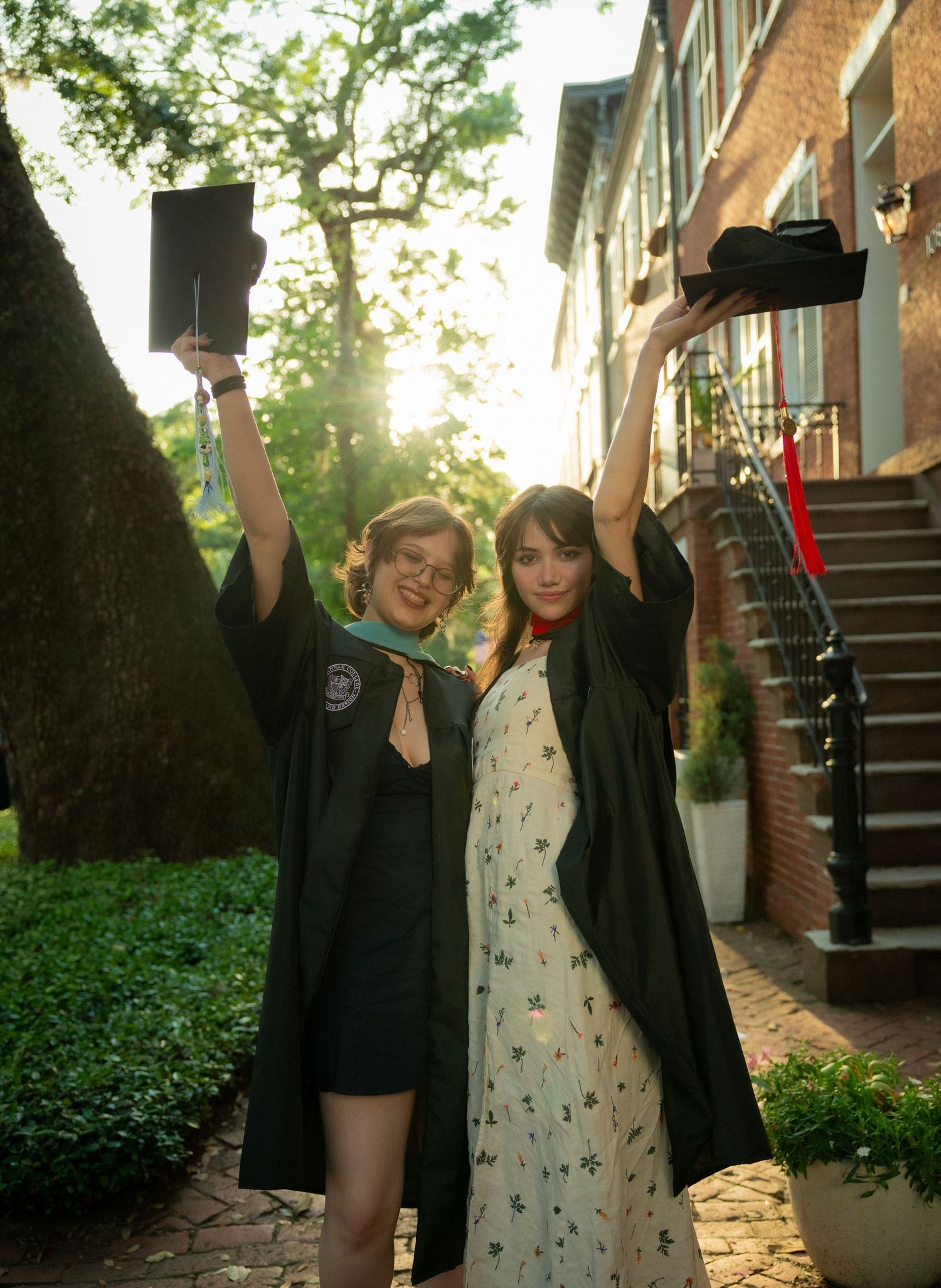 Two smiling graduates in black robes raise their caps outdoors near brick building in golden sunlight.