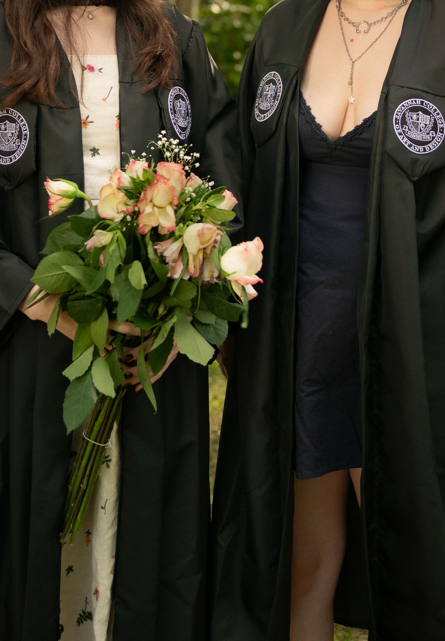 Two graduates in black SCAD robes hold bouquet of pink roses in outdoor garden setting.