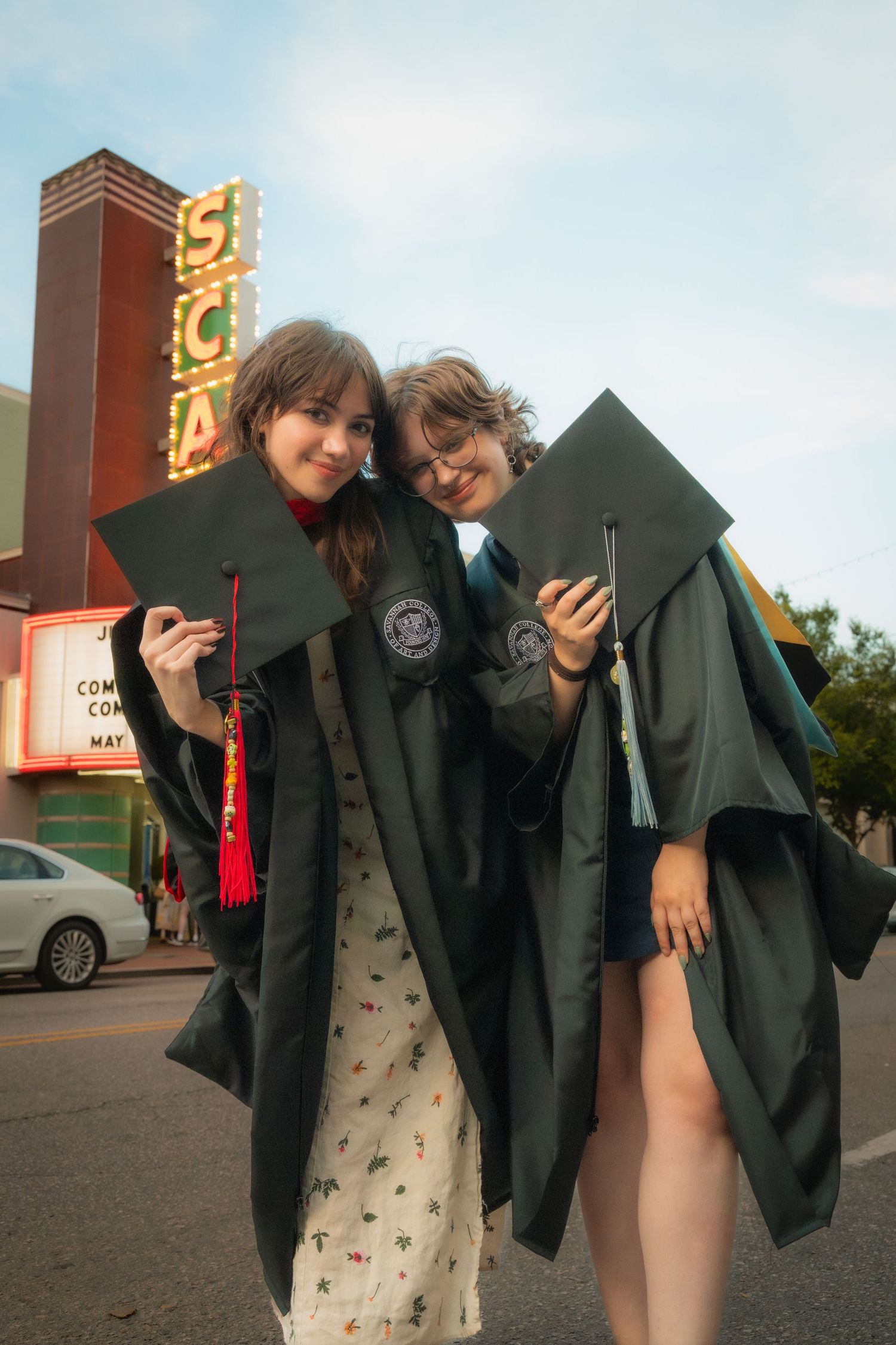 Two smiling female graduates in green robes pose with caps near vintage theater marquee sign.