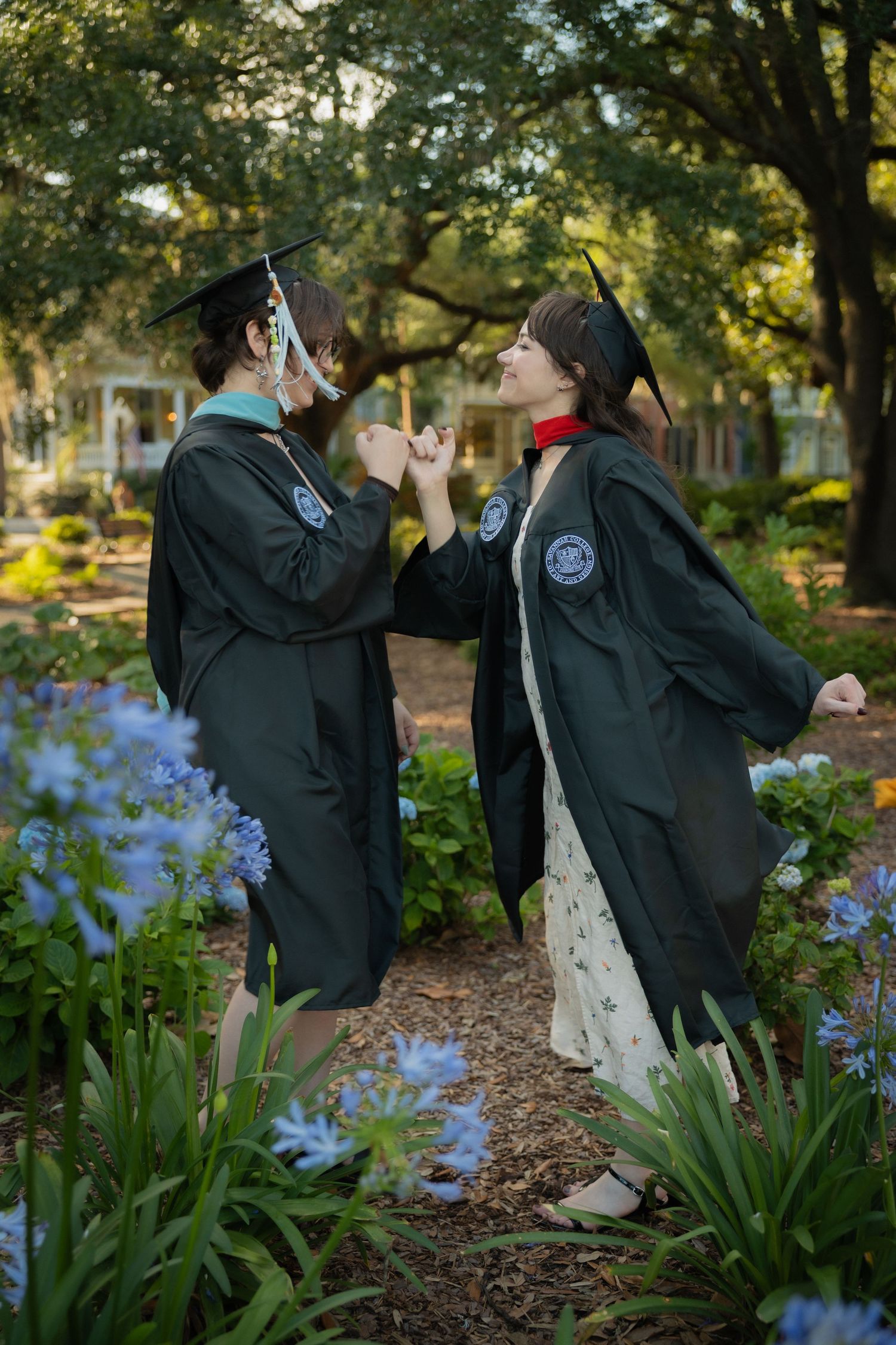 Two graduates in dark green caps and gowns hold hands and lean toward each other in a garden.