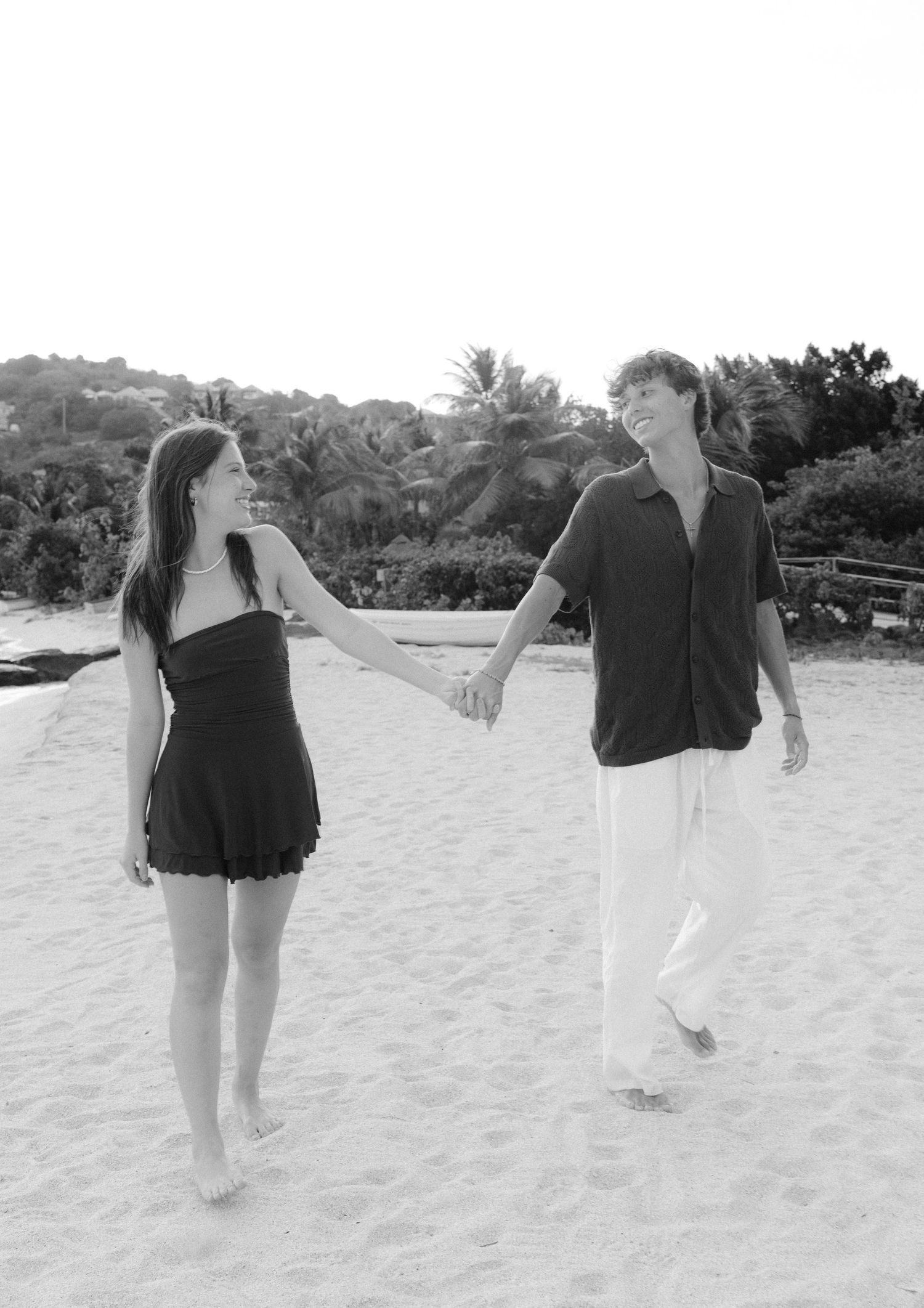 A young couple holds hands and walks barefoot on a sandy tropical beach, smiling at each other in black and white.