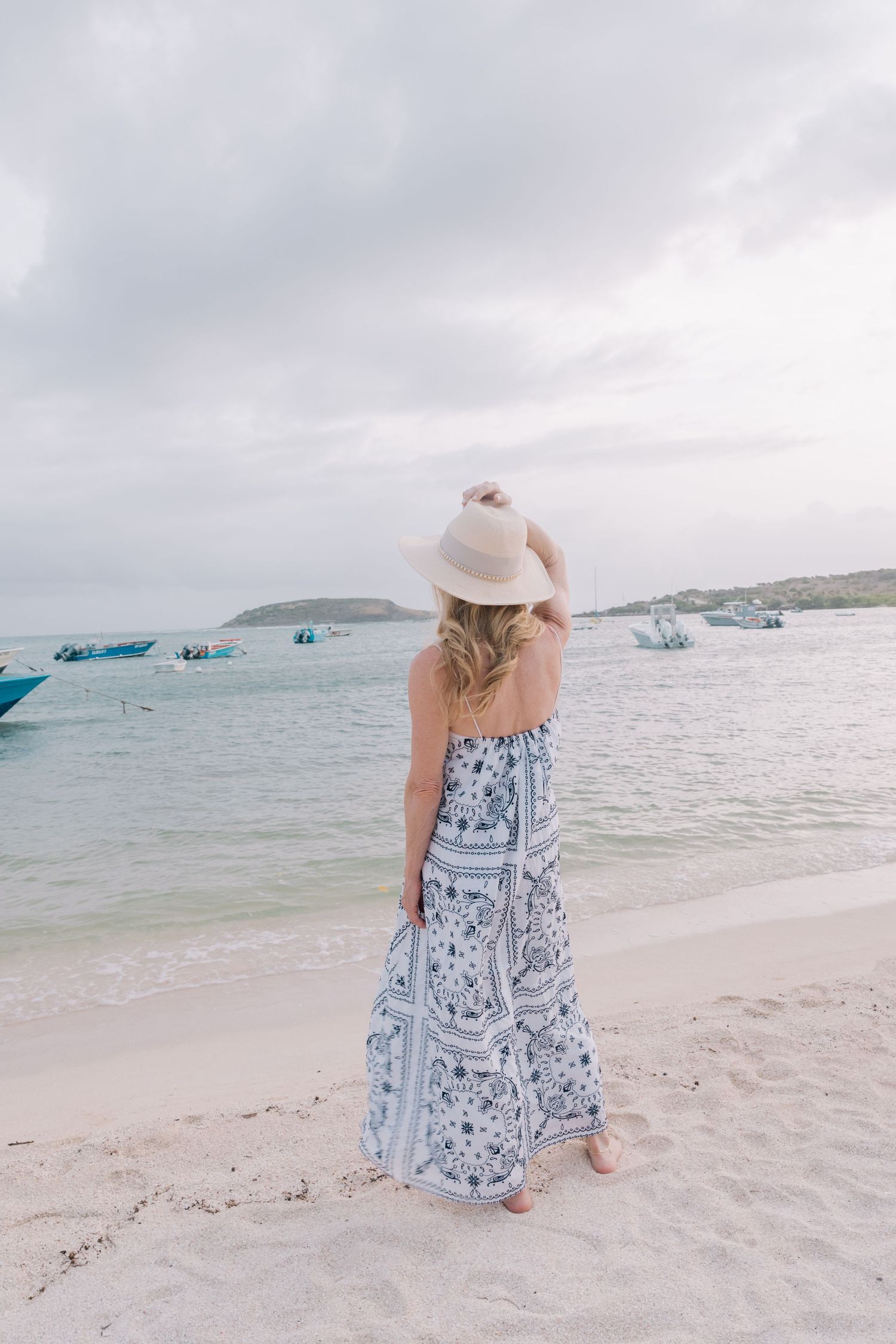 Woman in floral maxi dress and white sun hat standing on tropical beach with boats in calm turquoise water.