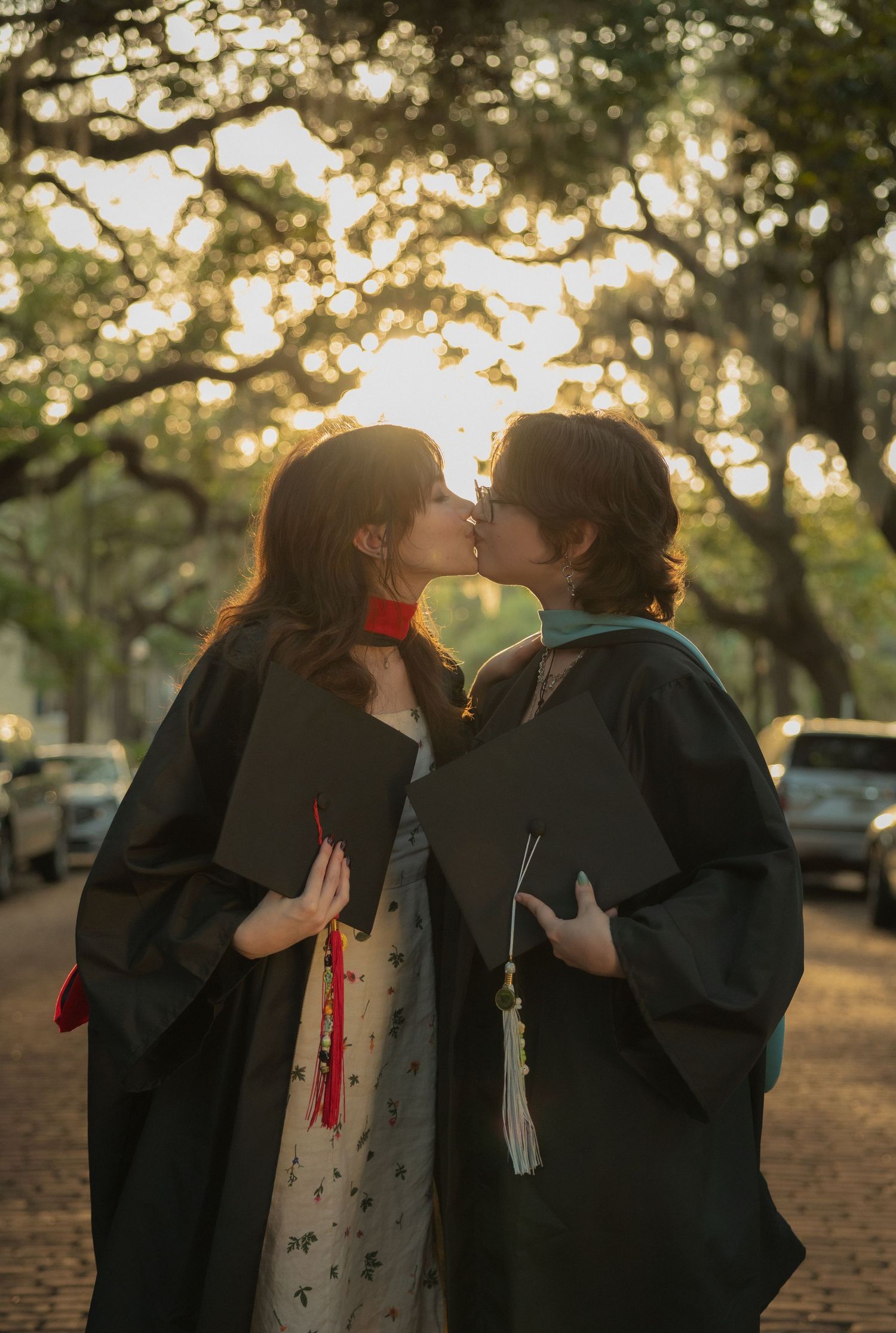 Couple in graduation gowns kiss on cobblestone path at sunset.