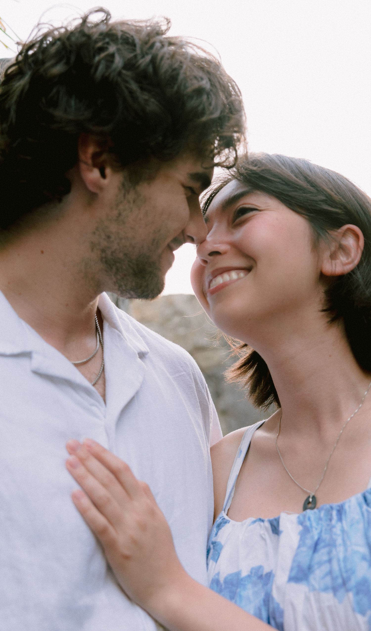 A smiling couple gazes lovingly at each other, nearly touching noses, in a tender romantic outdoor moment.